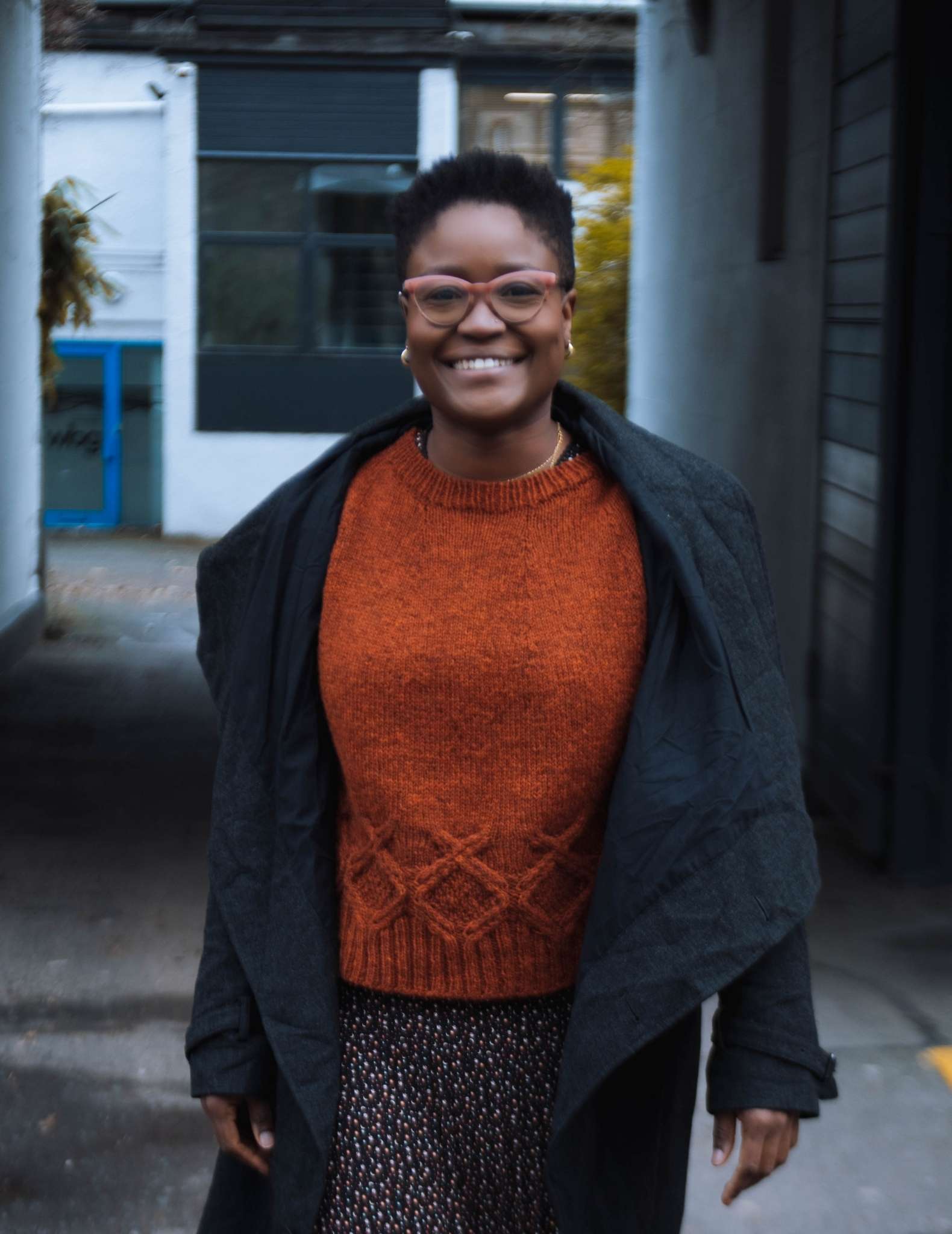 A black woman with short hair walks towards the camera wearing an orange cabled sweater, patterned skirt and a long dark jacket that hangs open.