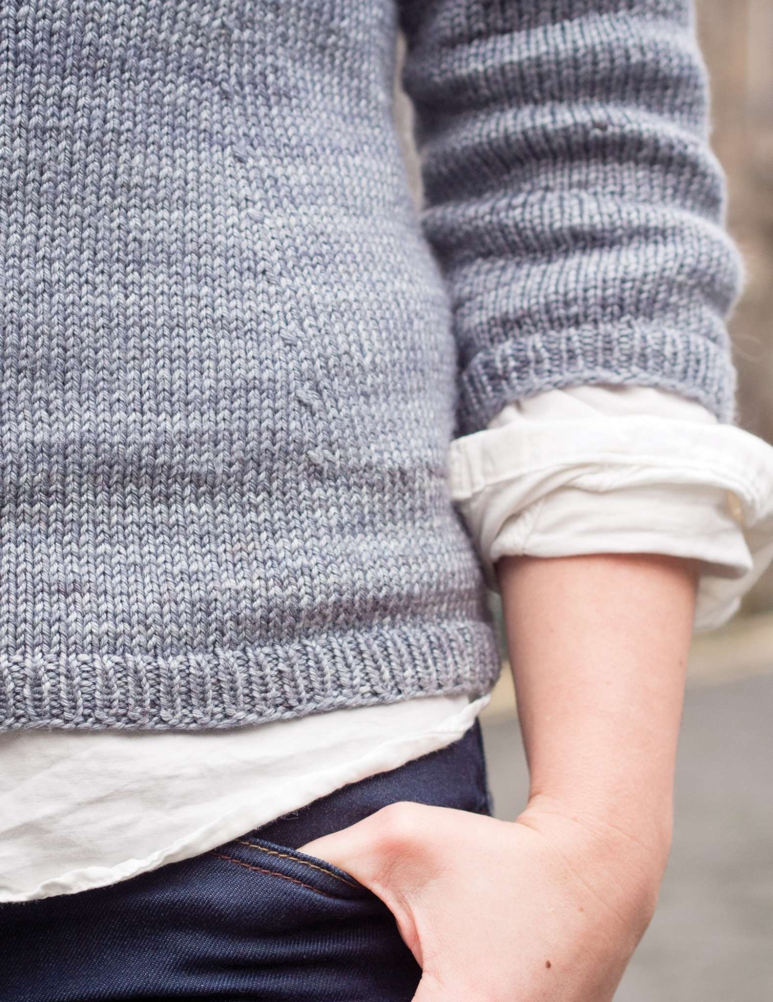 un primer plano de la parte inferior de un suéter gris, con un brazo colgando hacia un lado con la mano apoyada en la parte superior de un bolsillo de los vaqueros. Los dobladillos de una camisa blanca se ven en los bordes del dobladillo y del puño.