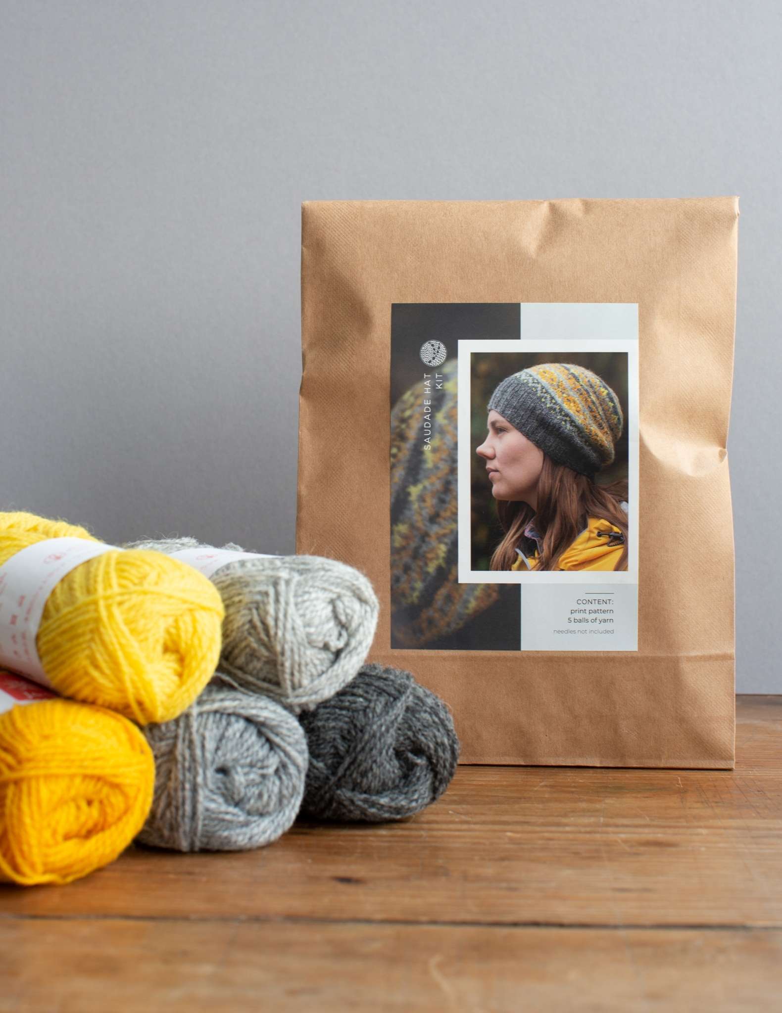 A hat knitting kit in a paper bag, sitting beside 5 balls of yarn in yellow and greys.
