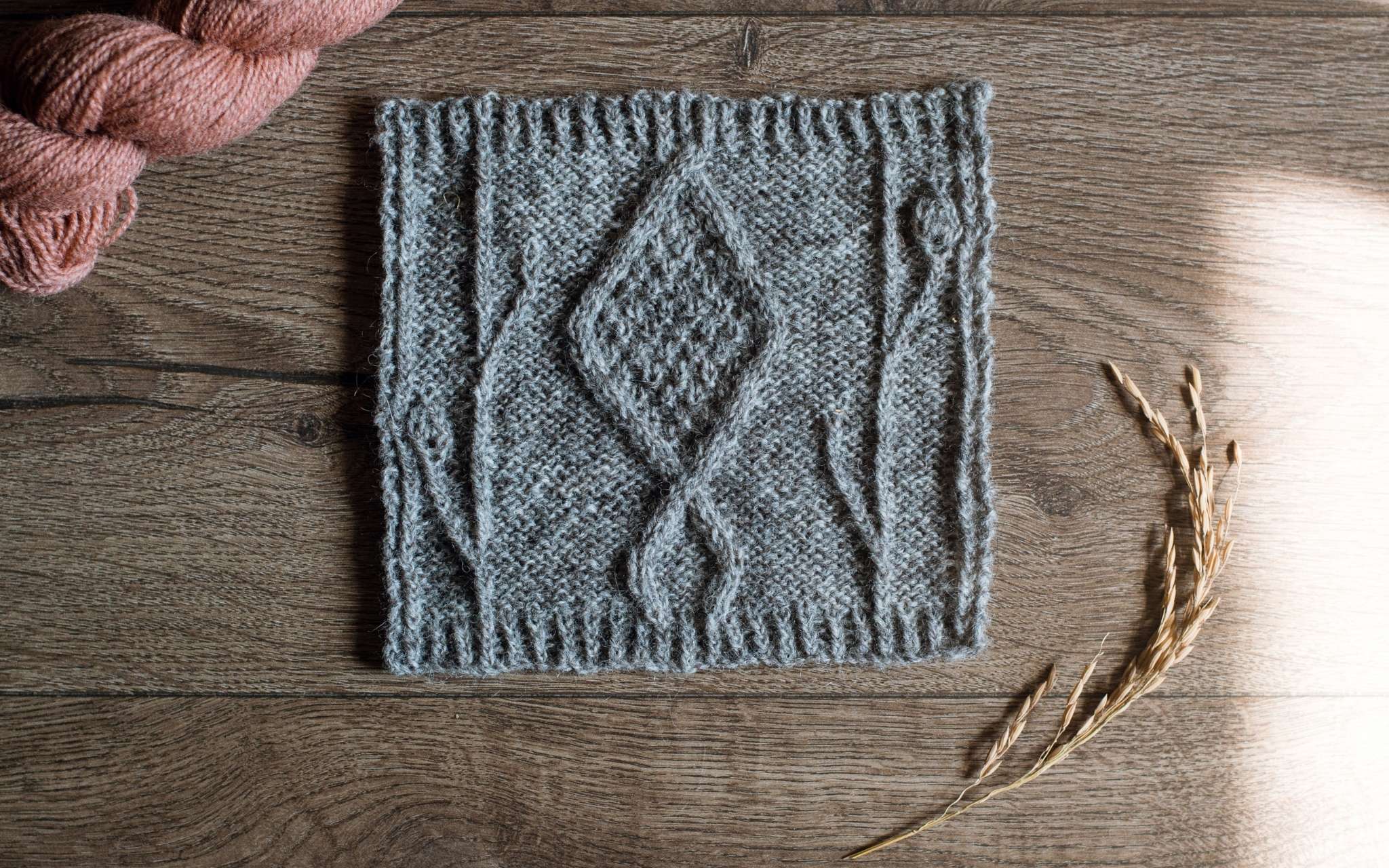 A swatch of cabled knitting in grey laid flat on a wooden surface. There are some dried stems, scissors and a bottle of pints to the side.