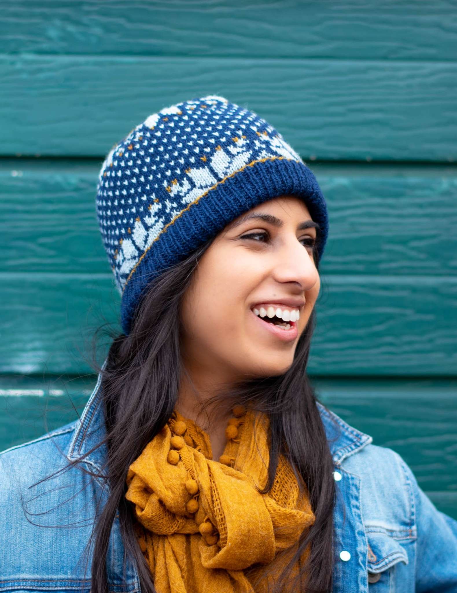 a woman with dark hair wears a dark blue and white colourwork hat, a yellow scarf and open denim jacket