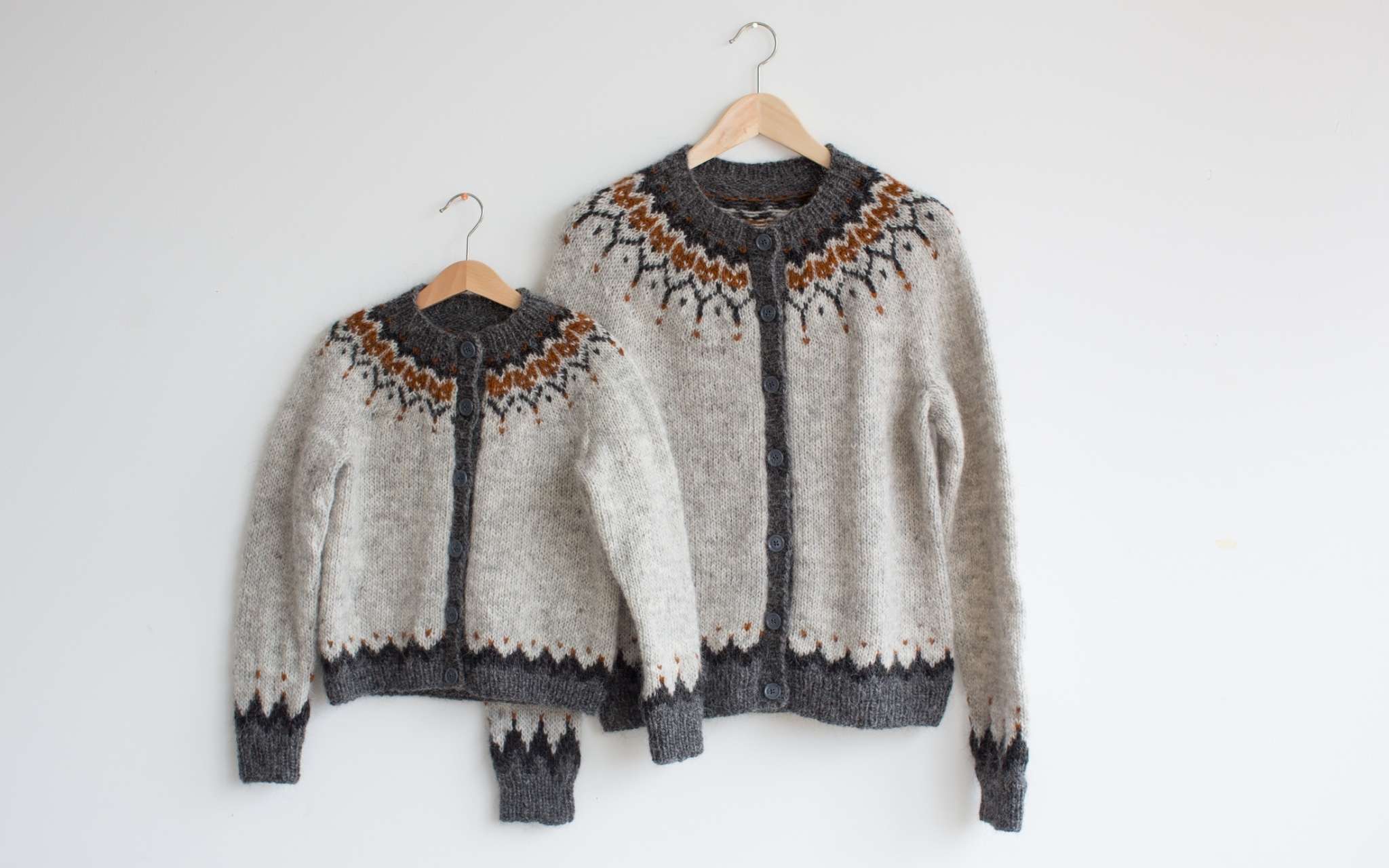 Matching adult and kid sweaters hang on wooden clothes hangers, overlapping against a wall. Both sweaters are grey, with a darker grey and orange colourwork yoke pattern.