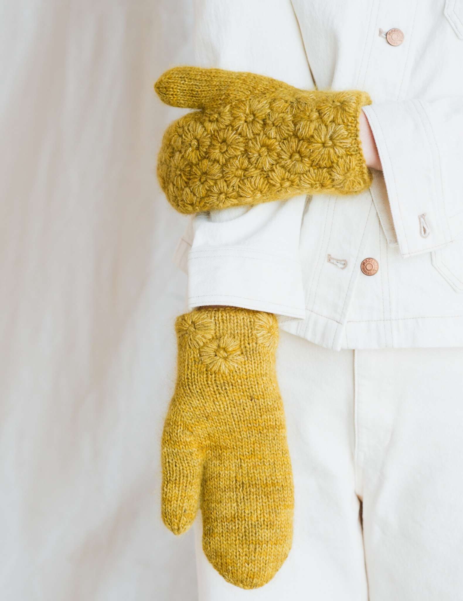A pair of mittens are worn by a model wearing white, one arm is behind held straight down with palm side up, the other hand is resting on the straight arm, showing the detailed back of the yellow mitten