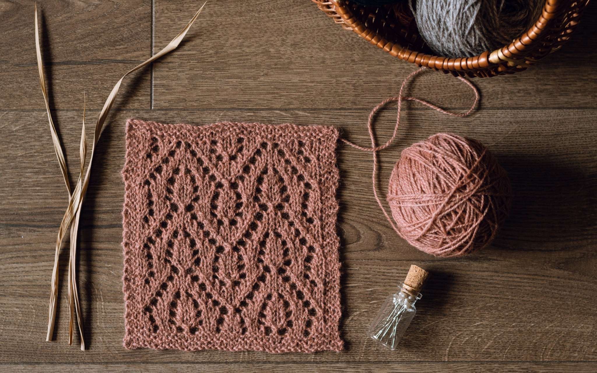A swatch of lace in coral laid flat on a wooden surface. There are some dried stems, scissors and a bottle of pints to the side.