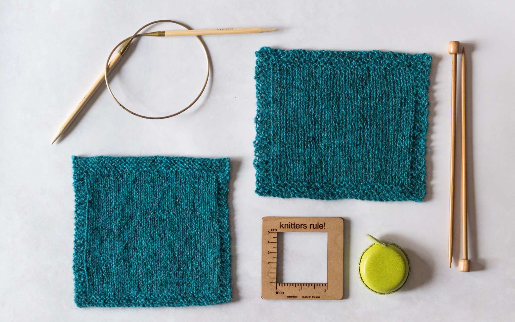 Two dark teal swatches in stocking stitch lie on a flat pale surface. The swatch in the bottom left of the image is smaller and more tightly knit, and the swatch in the top right of the image is larger and more loosely knit. Alongside the swatches lie a curled up circular needle, a pair of straight wooden needles, a lime green measuring tape and a wooden square measuring tool.