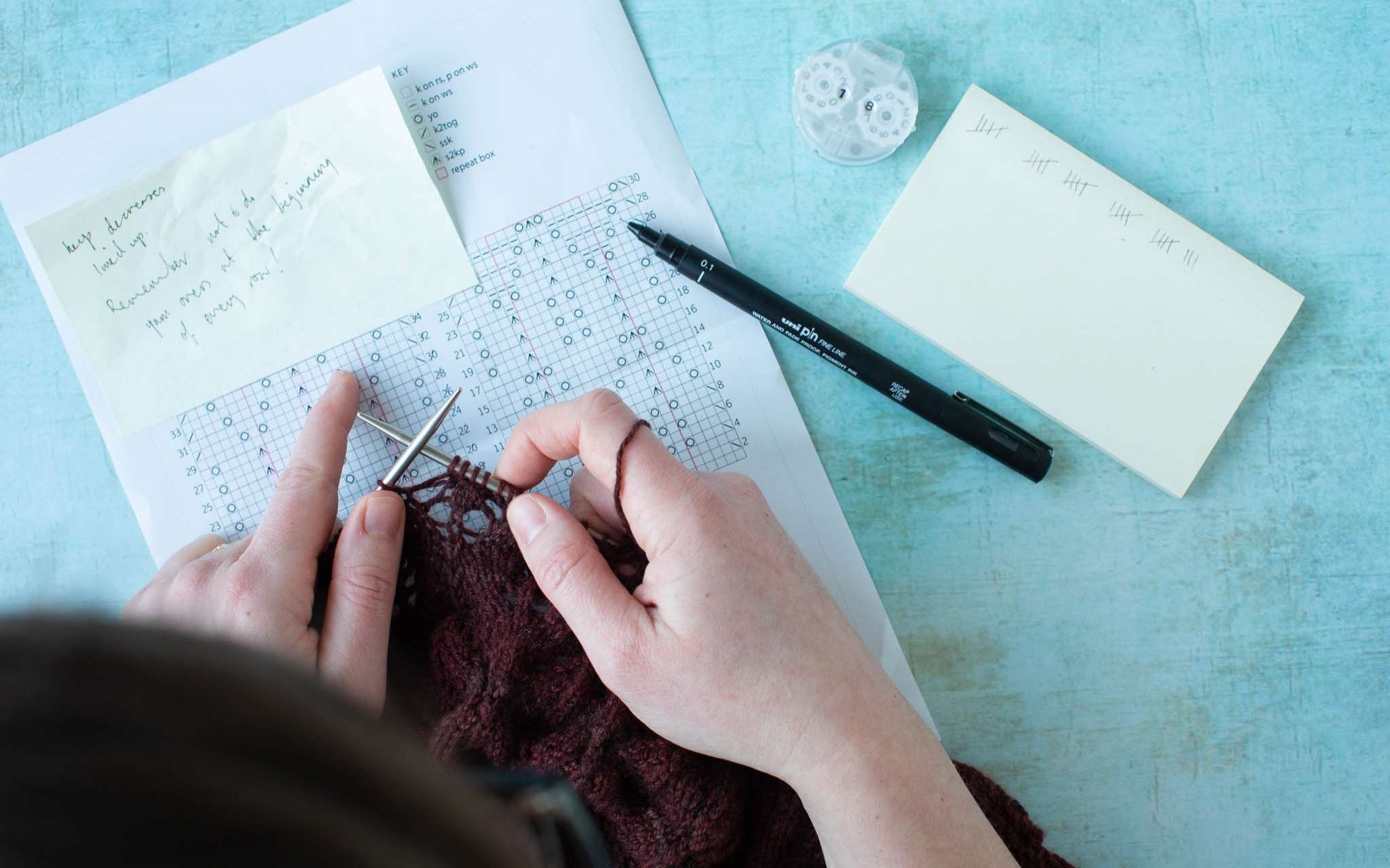 Hands are working on a brown lace knitting project. There is a paper pattern with handwritten notes on a blue surface, a pen, a knitting counter, and post it notes.