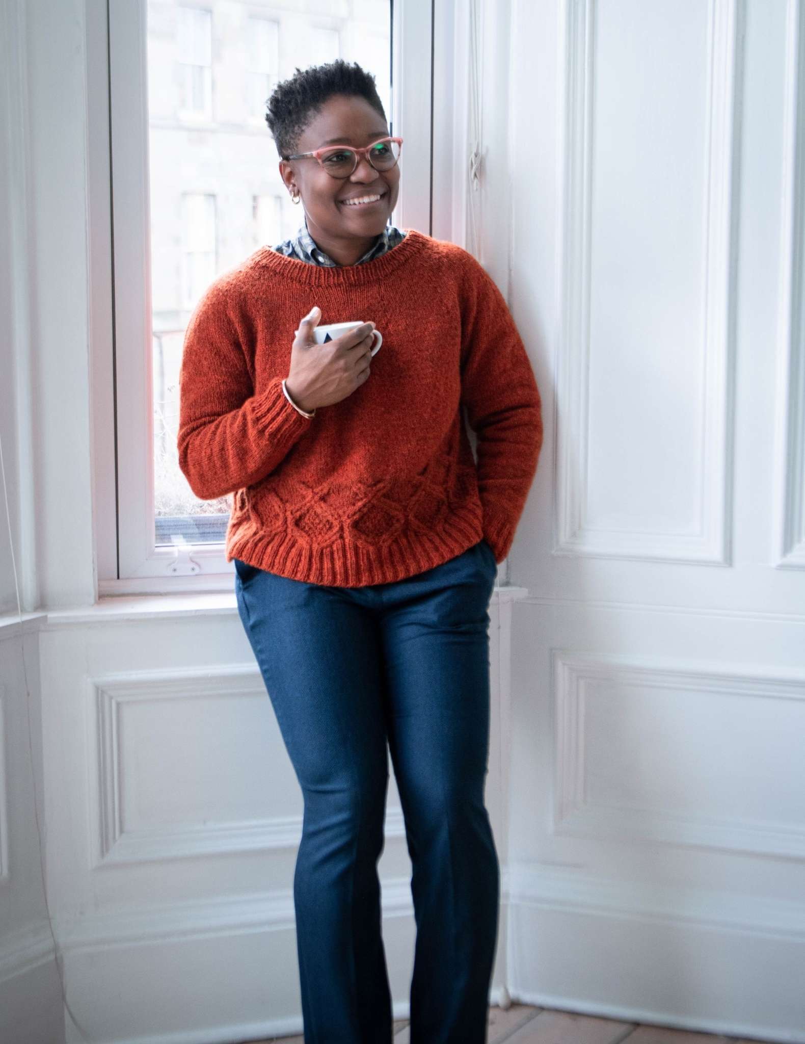A black woman relaxes against a white indoor wall, wearing an orange cabled sweater and jeans and holding a mug in her right hand.