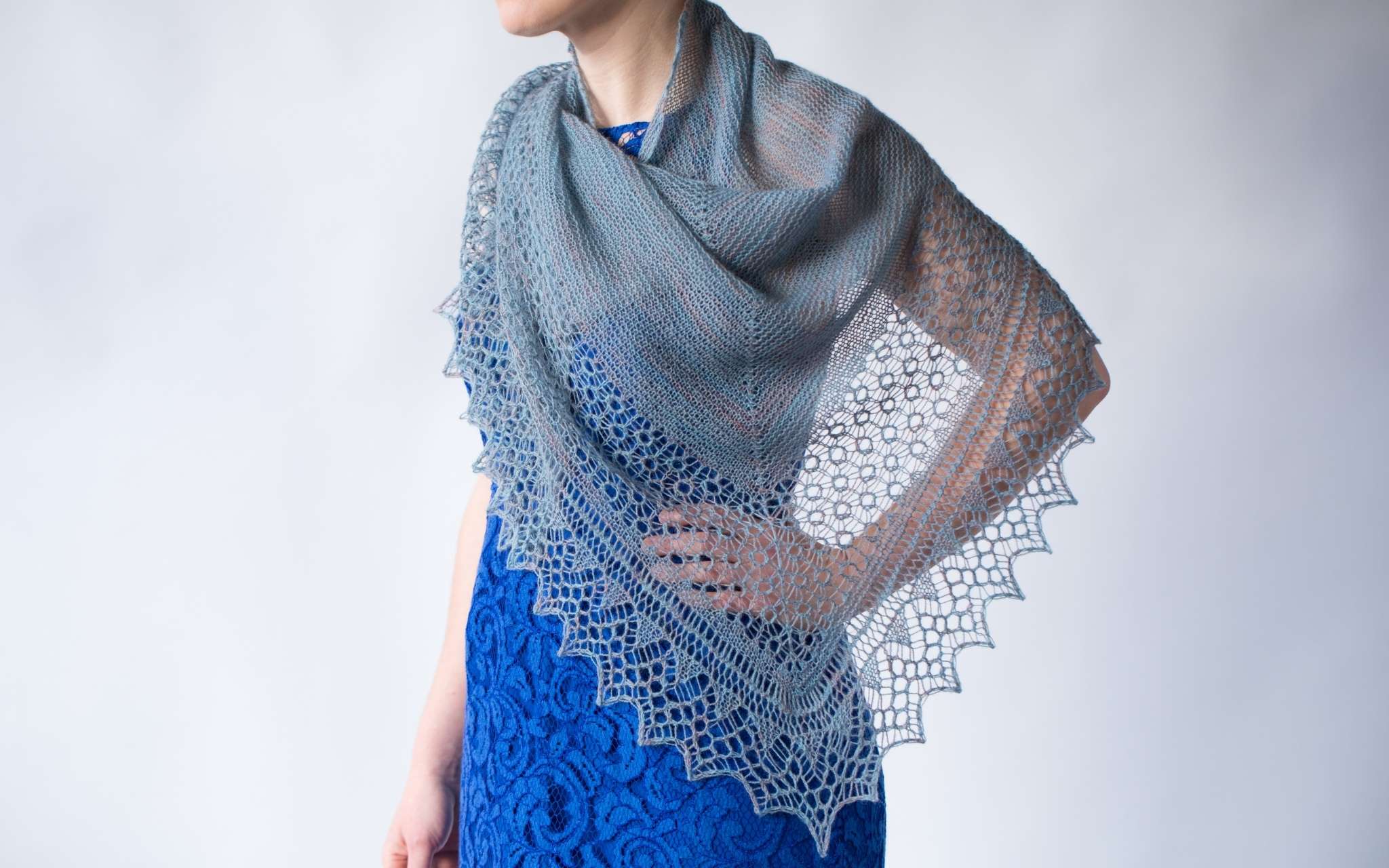 A close up of a delicate pale grey shawl, worn by a model wearing a blue dress. The model's head is not visible, but they are standing in front of a white background with their left hand resting on their hip.