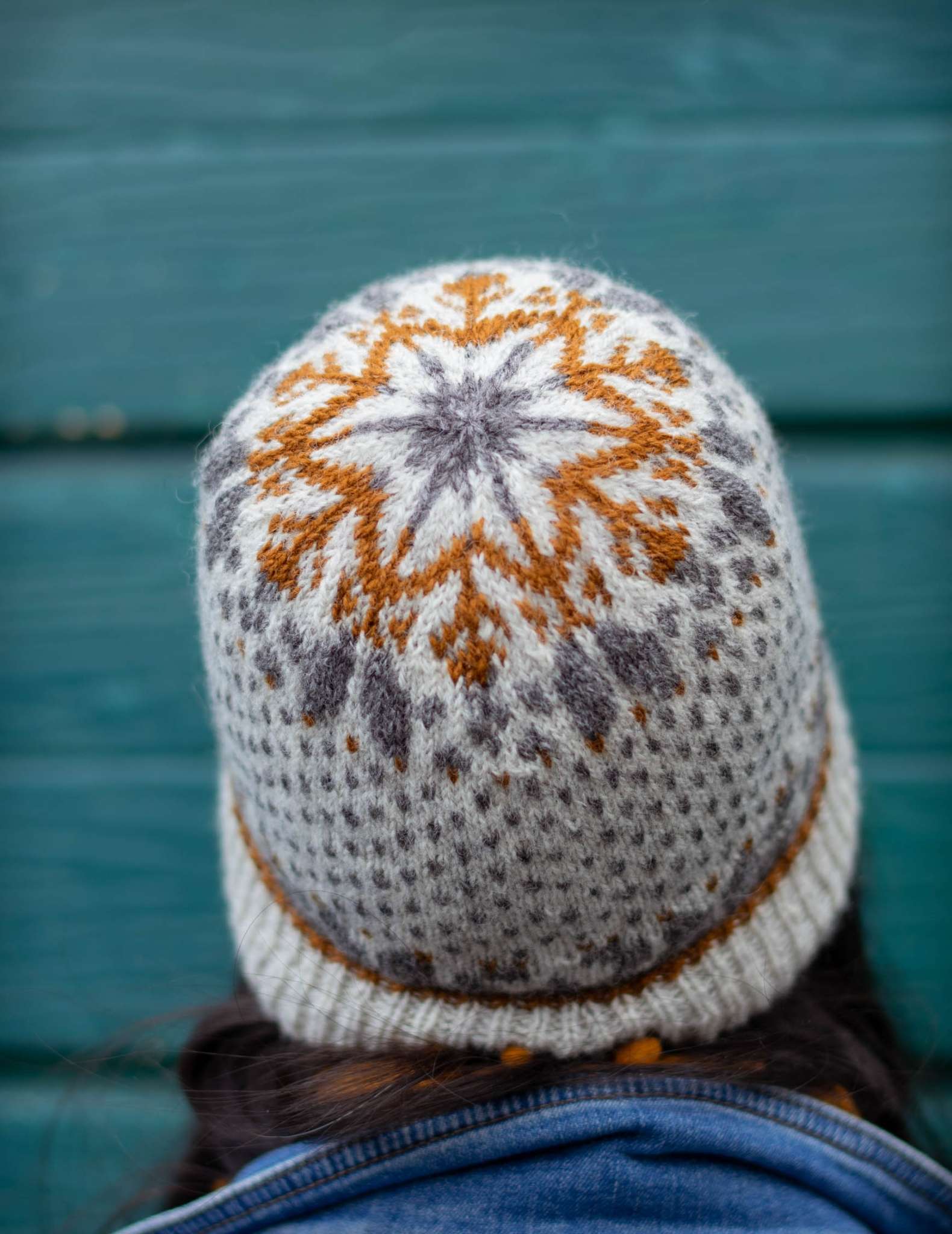 a model wears a cream, brown and grey colourwork hat with snowflake detail on the crown. Only the back of the head is visible.