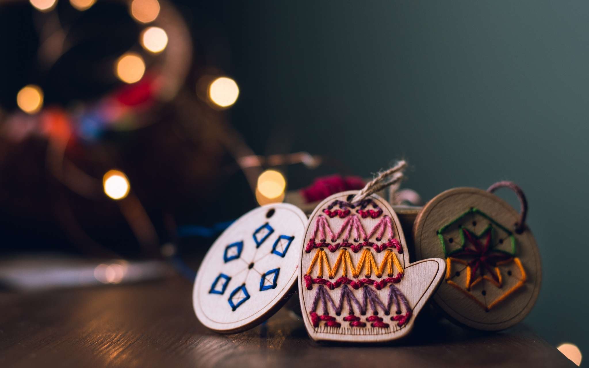 Wooden hanging ornaments that have been hand stitched in bright colours are gathered on a wooden surface, with fairy lights and a tree blurred behind.
