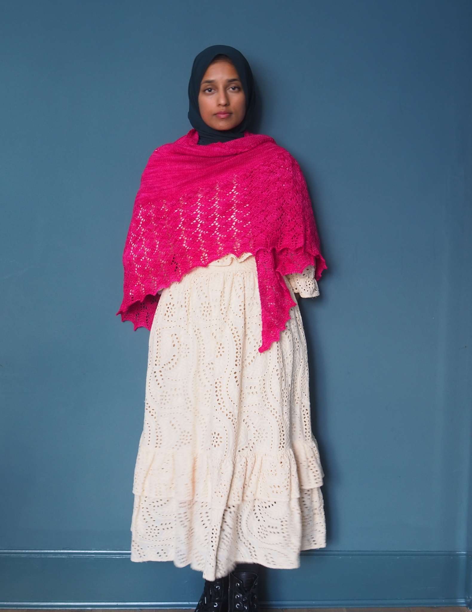 A brown woman in a hijab stands in front of a dark blue background. She is wearing a floaty cream dress with a large bright pink shawl wrapped around her shoulders and chest.