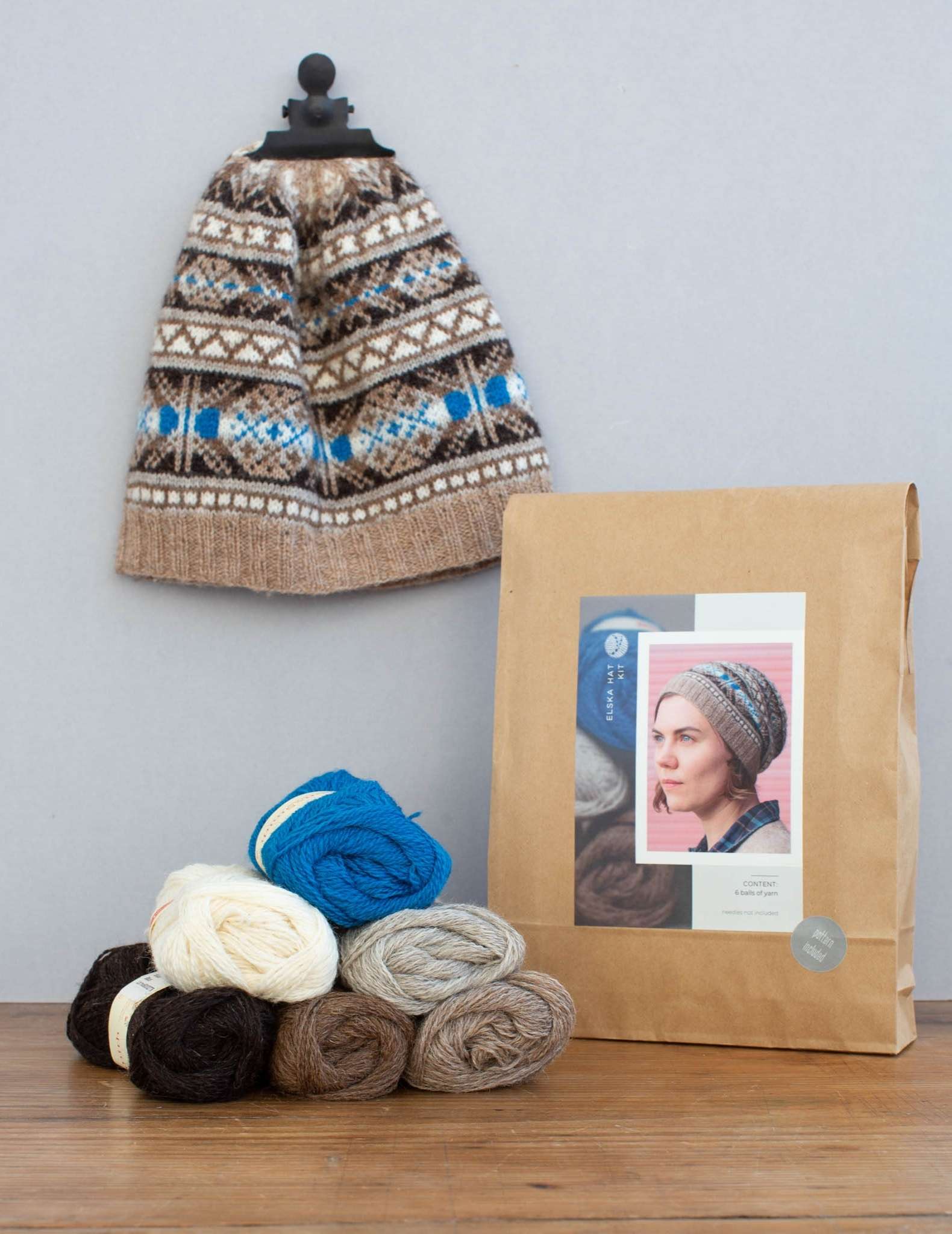 A hat knitting kit in a paper bag, with the hat hanging from the wall behind by a clip and 6 balls of yarn piled in front in blues and neutral colours.