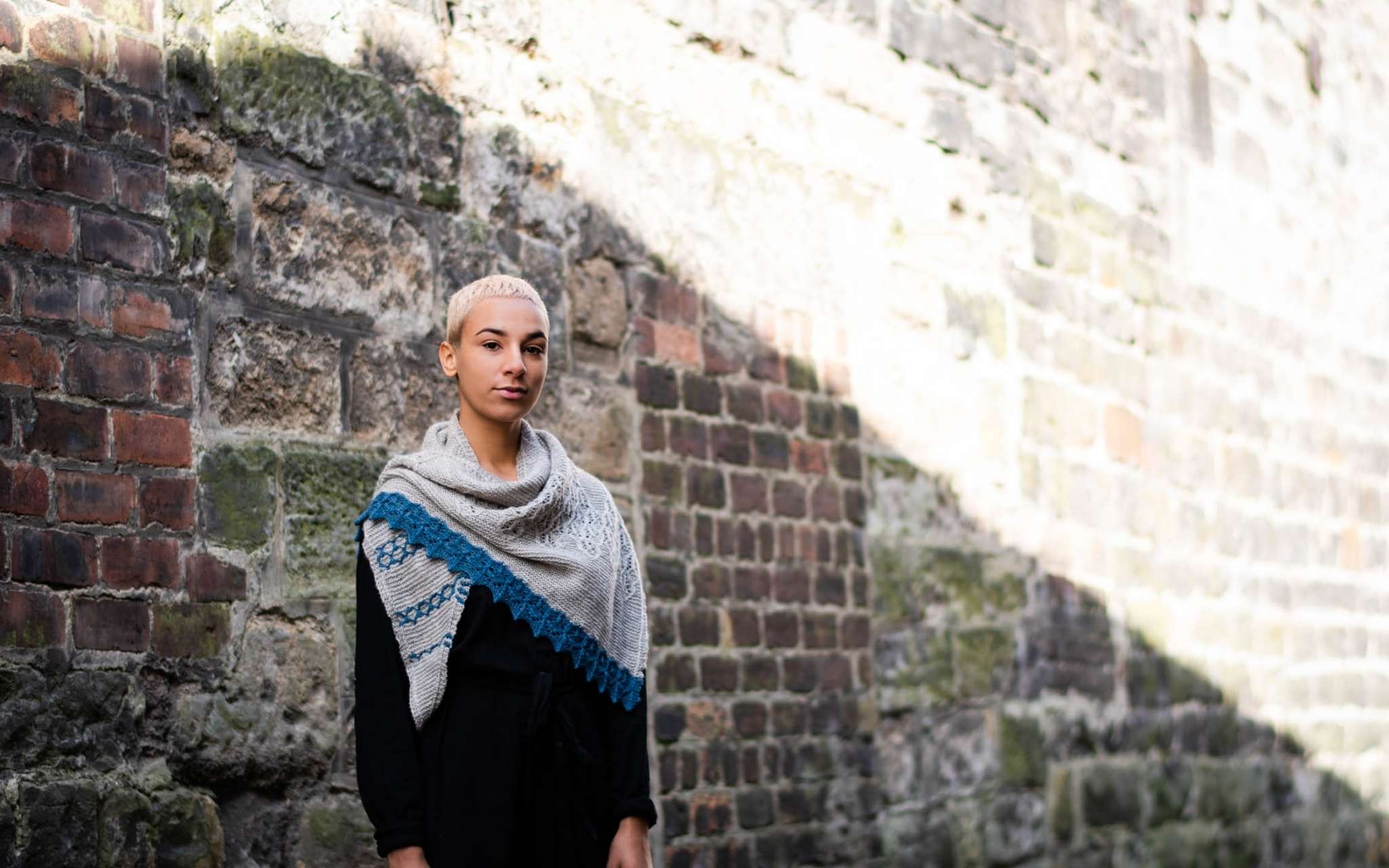 A model with short pale hair stands against a brick wall in the bright sunshine. They are wearing dark clothes, and a pale grey shawl with blue contrast edging is around their neck.