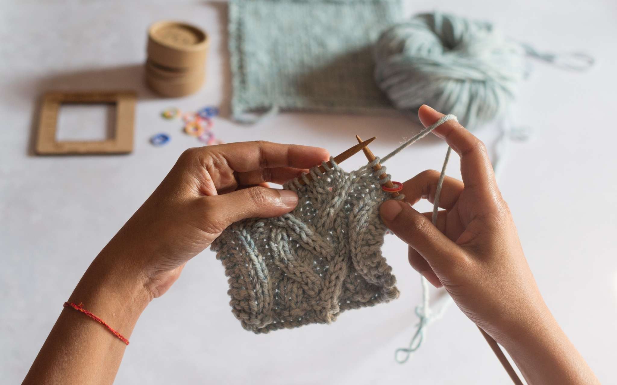 A woman of colour knits a pale blue cabled swatch on wooden needles. A completed swatch and measuring tools are on a flat surface at the top of the image.
