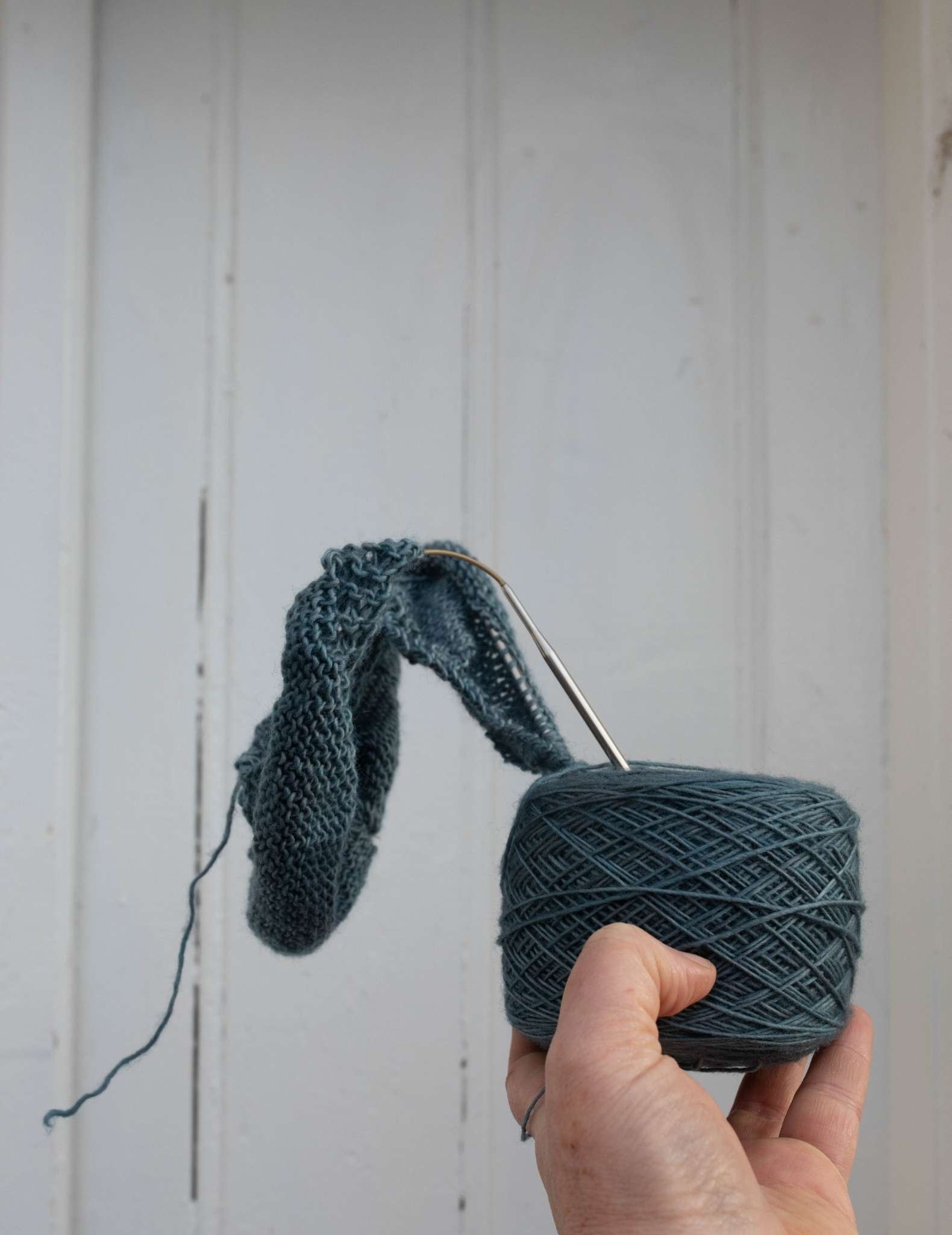 a white hand holds a ball of blue yarn with a project on the needles sticking out of the top, stretching away from the camera.