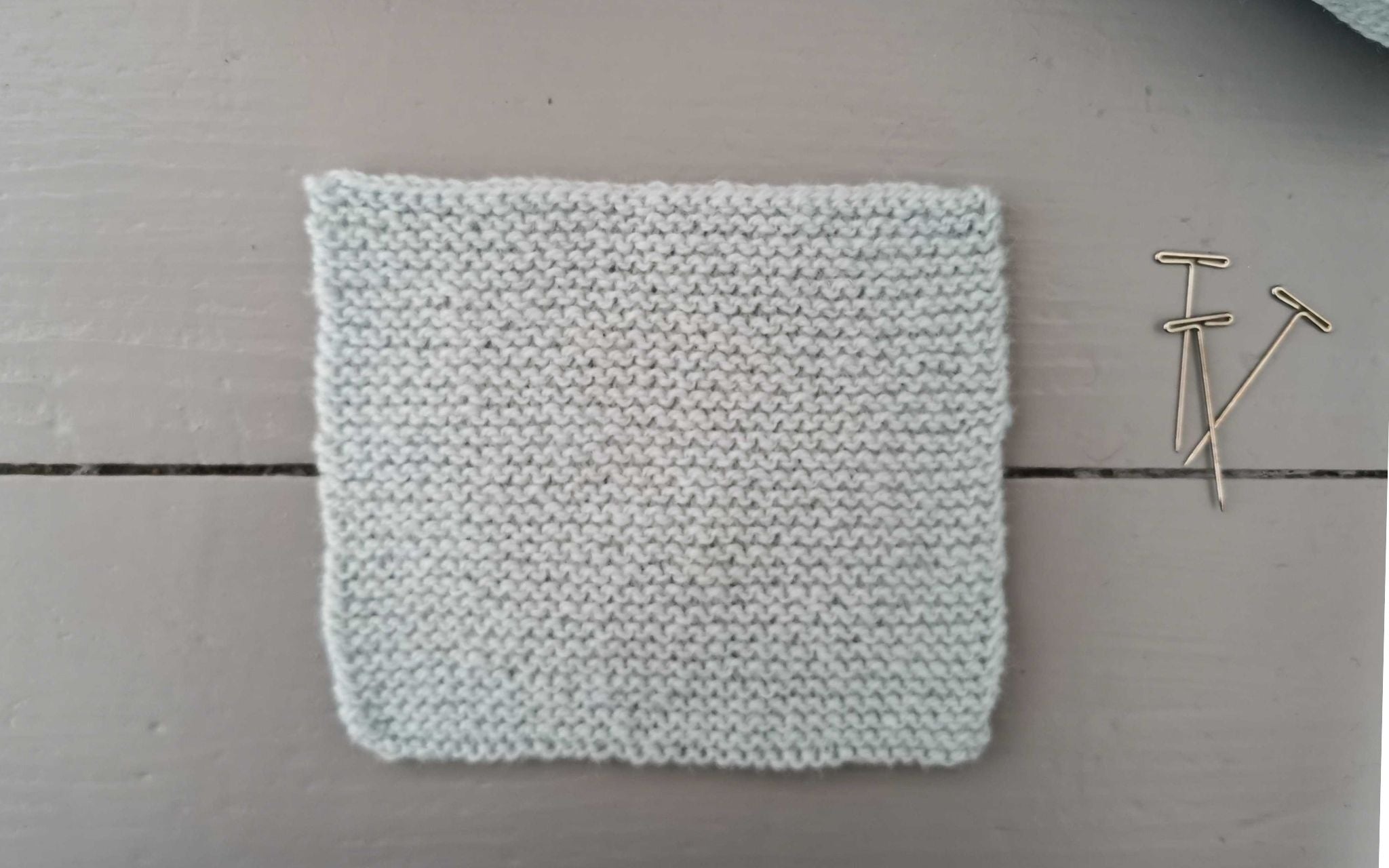 a square swatch of garter stitch in a pale blue yarn, with three t-pins at the side