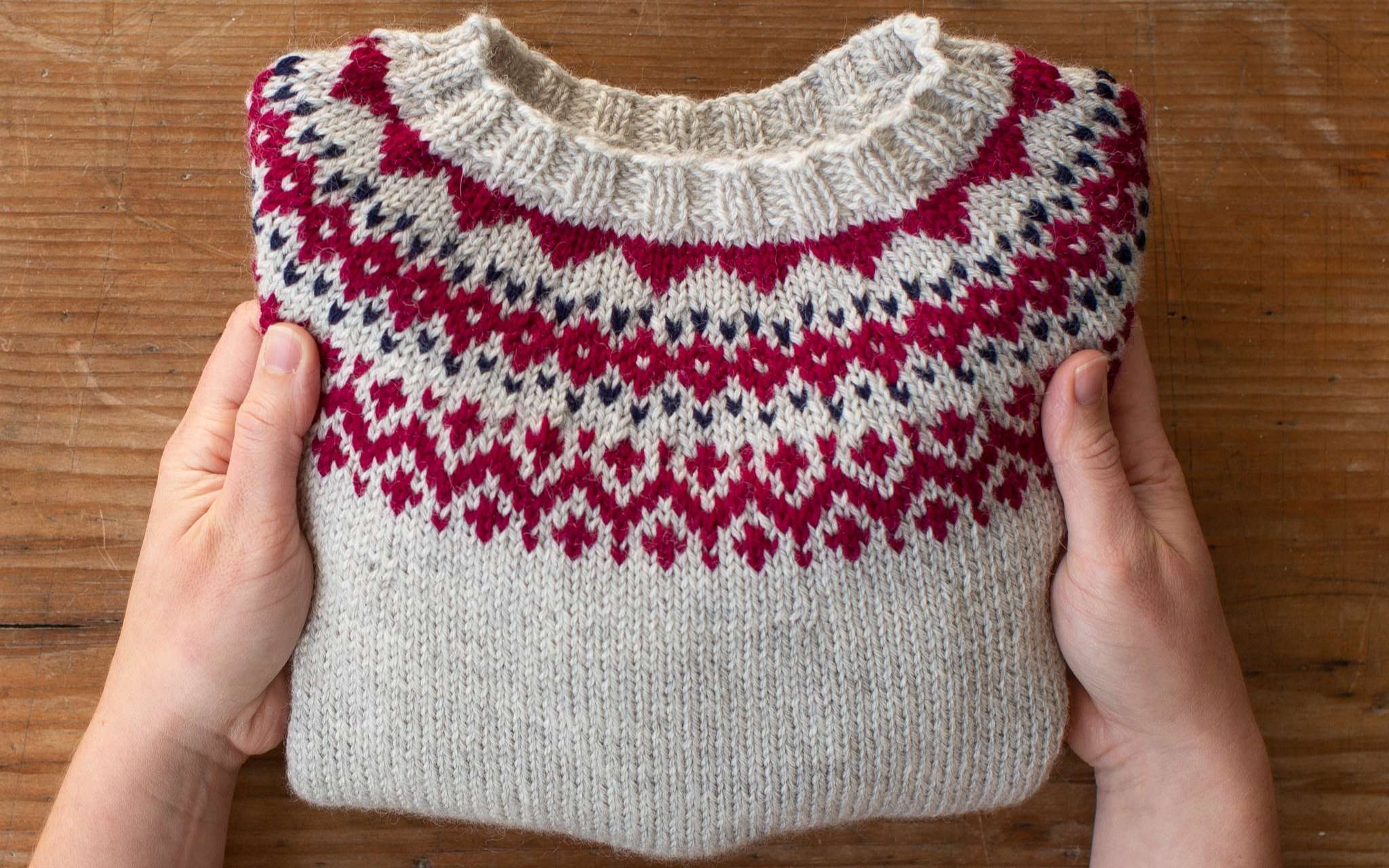 Two hands hold a folded sweater over a wooden surface. The sweater is pale grey with a pink and navy colourwork pattern around the yoke.
