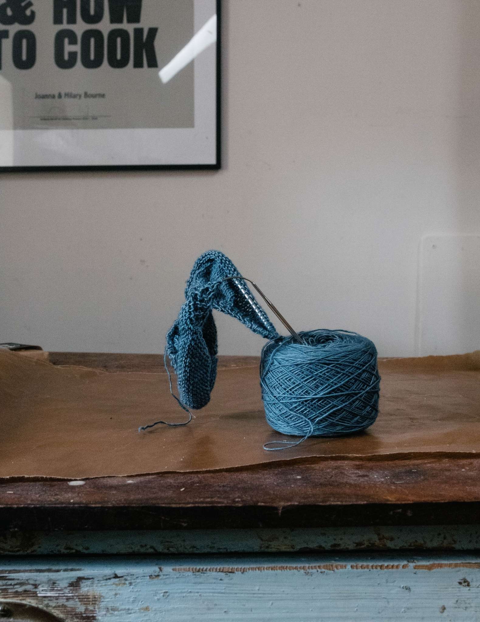 A ball of blue yarn with a project on the needles sticking out of the top of it, sitting on a wooden surface in front of a wall with an art print.