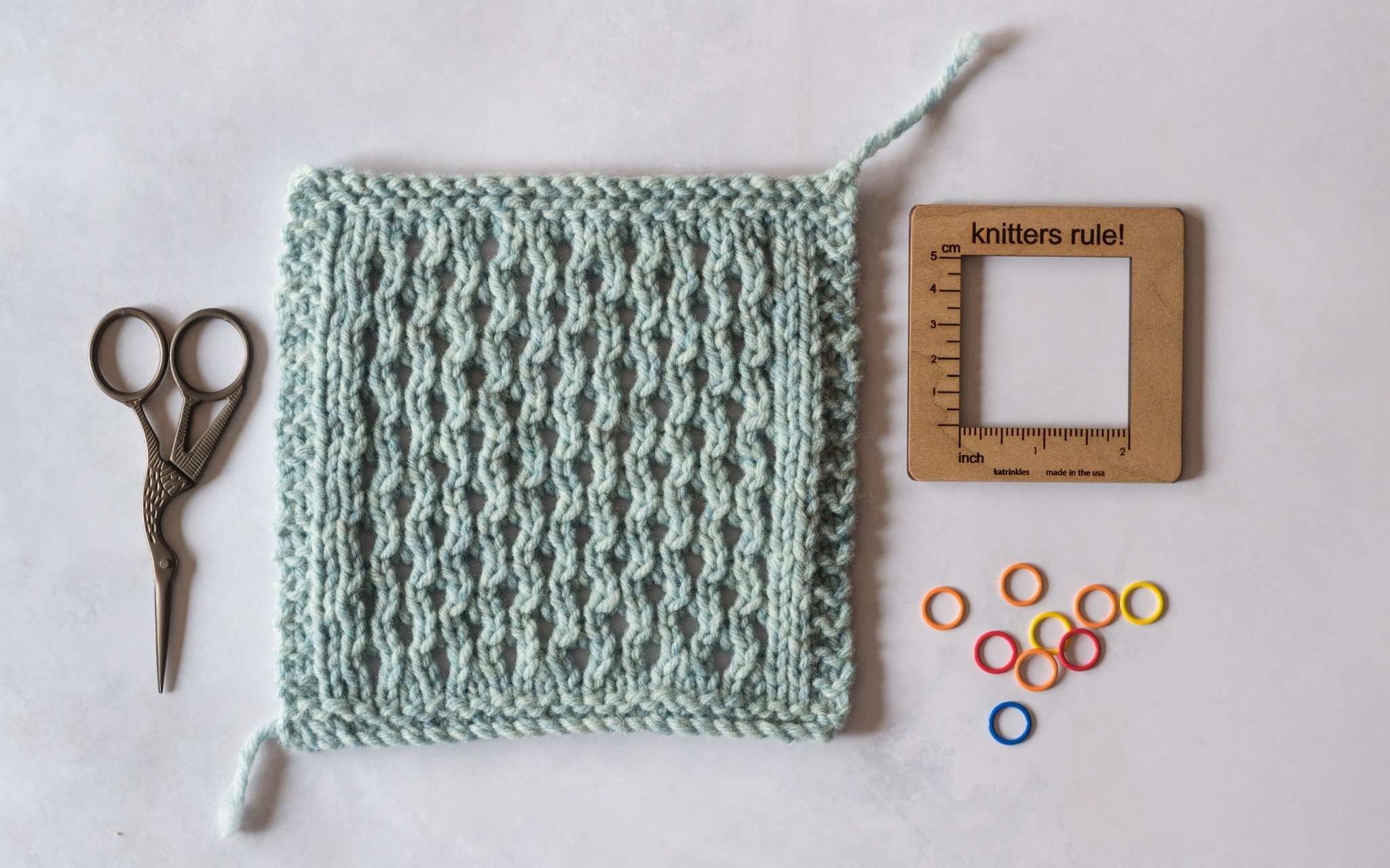 A pale blue cabled swatch lies on a flat surface. A small pair of scissors lie next to it on the left, and to the right is a square wooden swatch measuring tool and brightly coloured stitch markers.