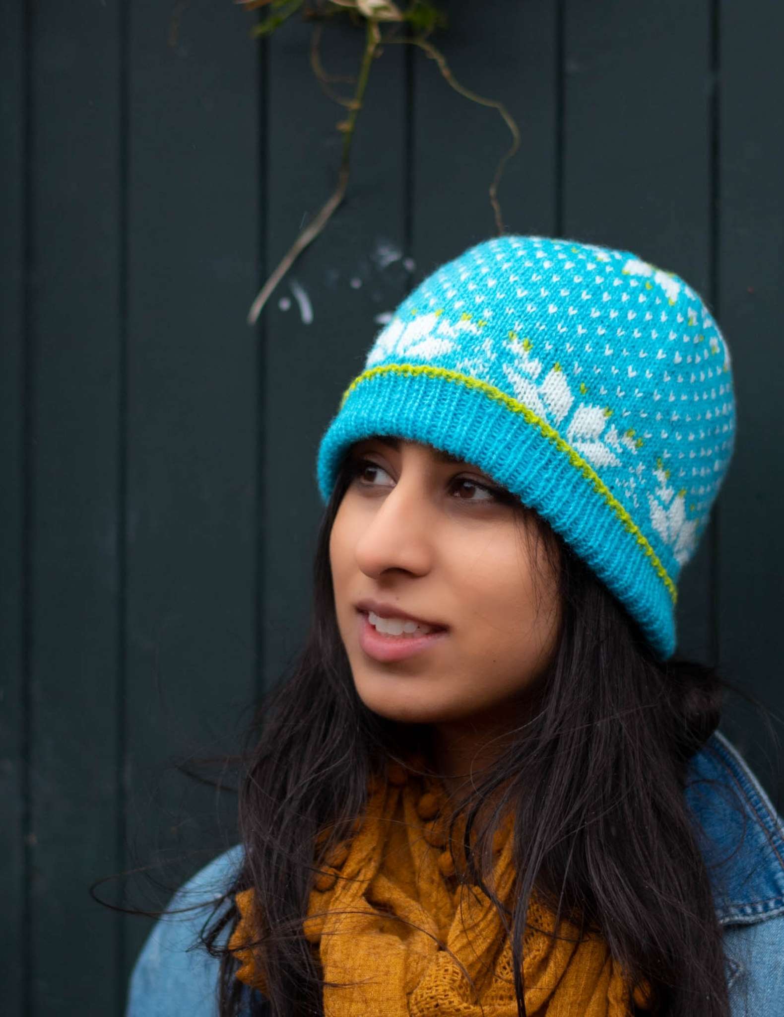 a model with dark hair wears a bright blue colourwork hat with snowflake detail. They are wearing a yellow scarf and looking slightly to the side.