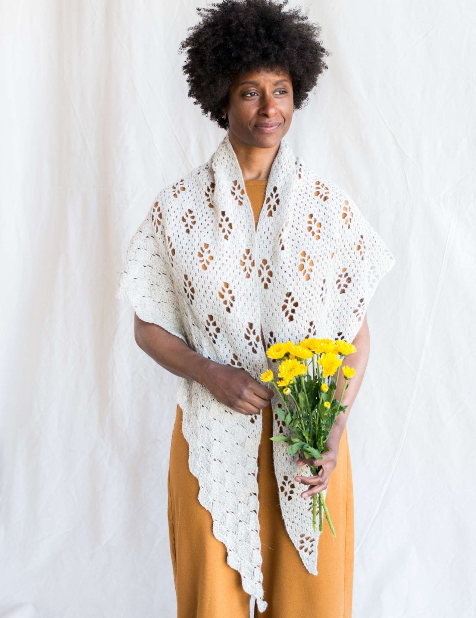 A large crocheted white shawl with lace details is worn by a black woman holding a bunch of yellow flowers. The model faces the camera with the corners of the shawl draped over each shoulder.