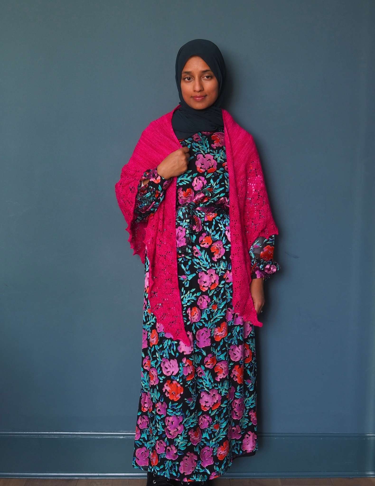 A brown woman in a hijab stands in front of a dark blue background. She is wearing a vivid floral print dress in black, pinks, orange and greens. She has a large bright pink shawl around her neck with the pointed ends hanging down at the front, and her right hand is raised and holding the straight edge of the shawl along her chest.