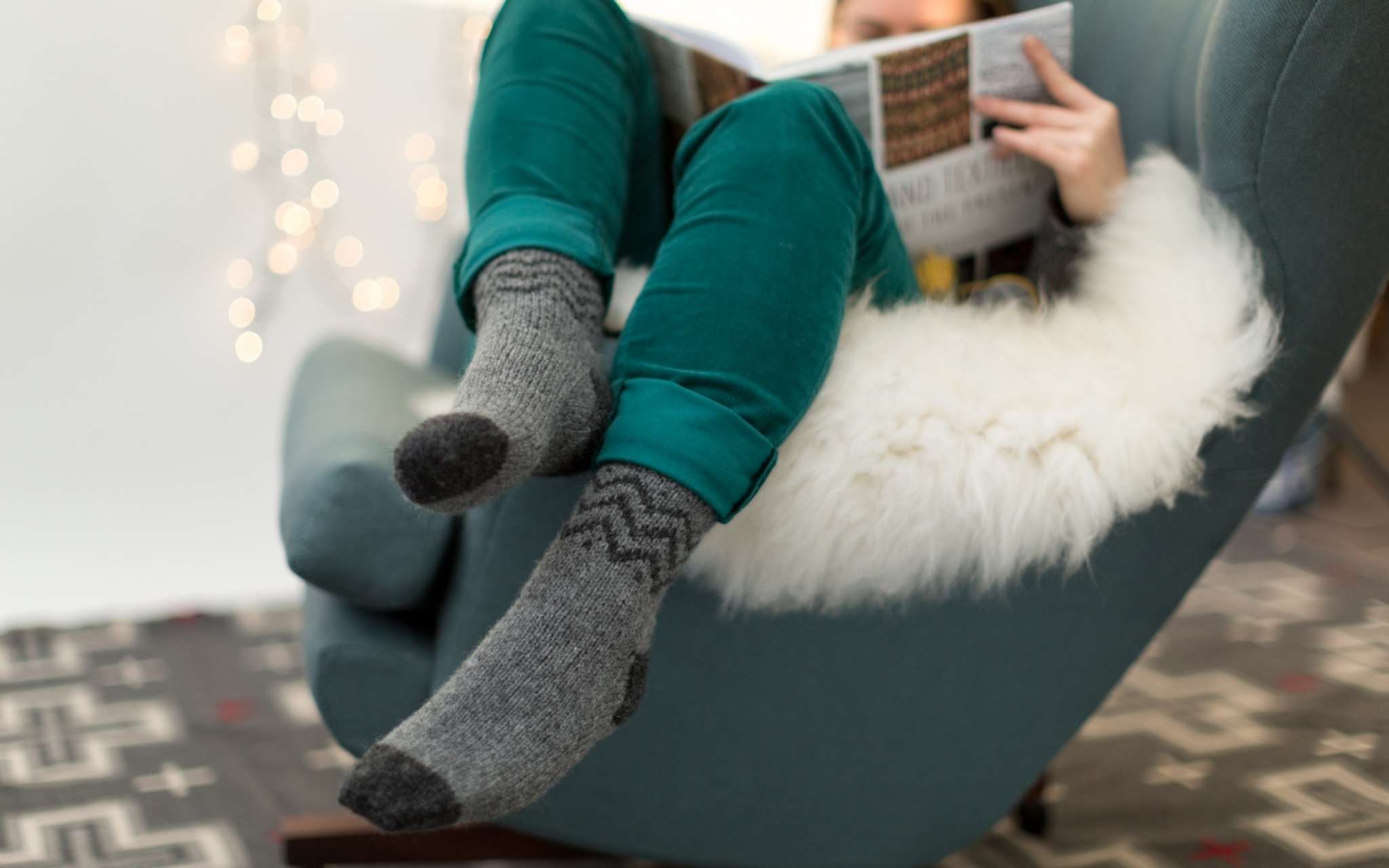 A model wears grey colourwork socks and teal trousers, while snuggled on an armchair holding a book. There are twinkling lights in the background and they sit on a fluffy white blanket.