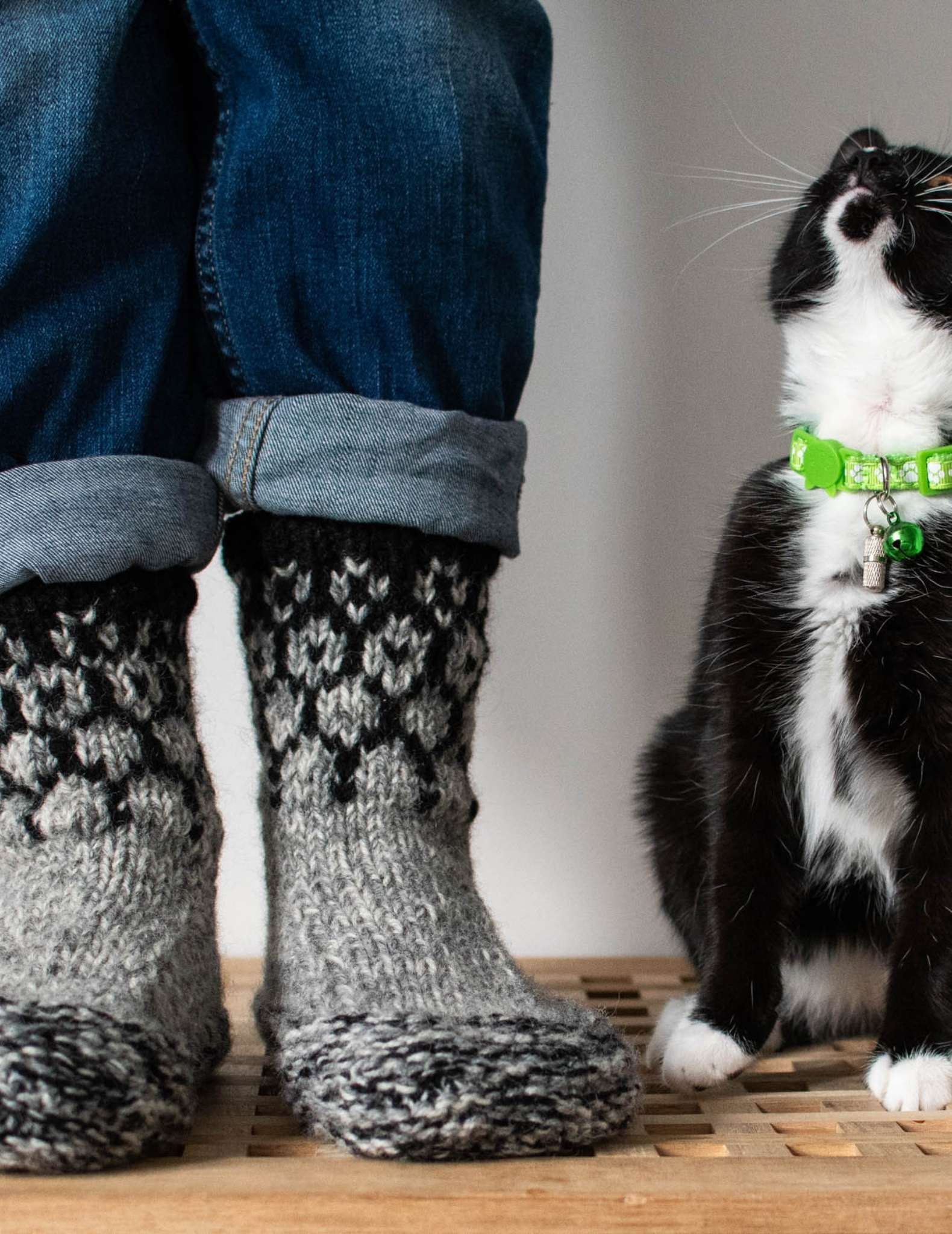 a model wears a pair of knitted slippers with colourwork detail on the cuff, they standing on a table with just their feet and cuffs of rolled up jeans showing. Next to them sits a black and white cat with green colour that looks up at the wearer.