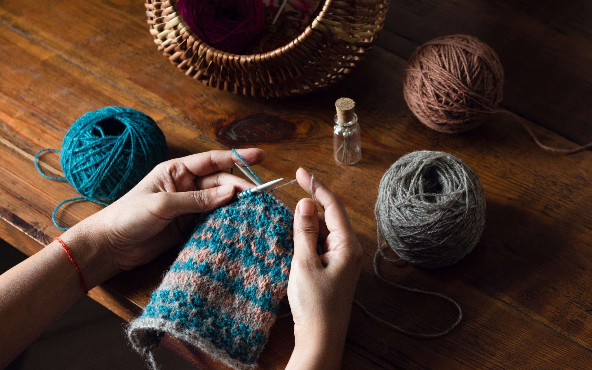 Hands knitting on a colourwork swatch in blue, coral and grey. The knitting is being held over a wooden table and there is a round wicker basket at the top of the photo and a bottle of pins.