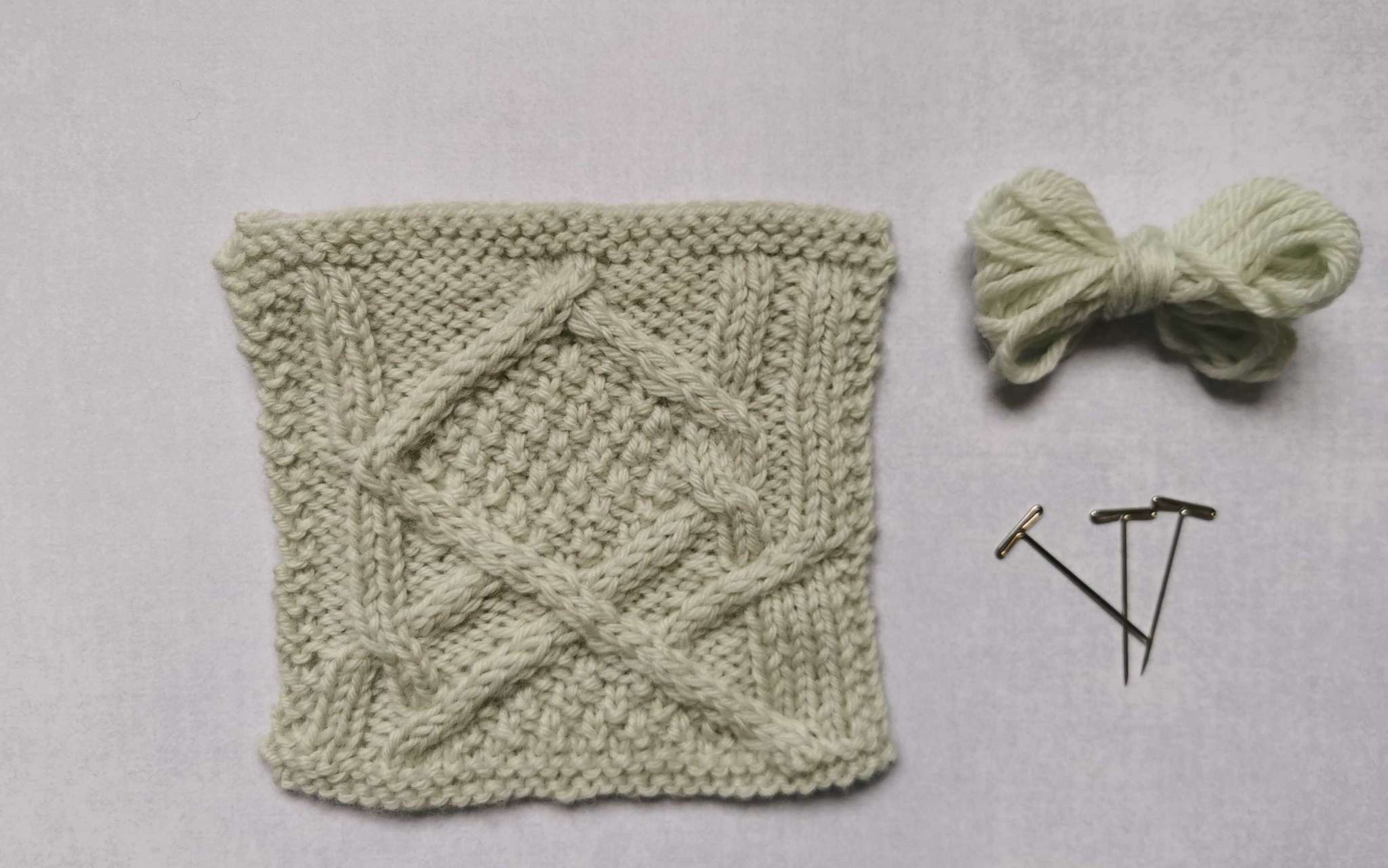 A square swatch of stocking stitch in pale cream, with some bundled up spare yarn and t-pins laying beside it to the right.