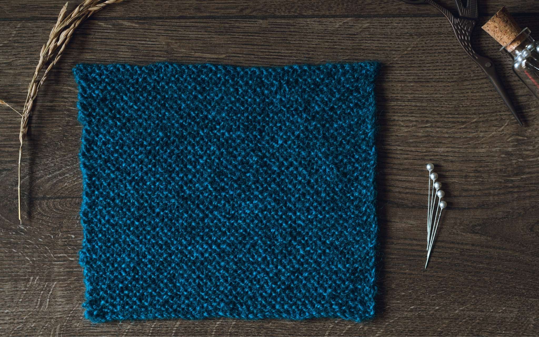 A swatch of garter stitch in dark blue laid flat on a wooden surface. There are some dried stems, scissors and a bottle of pints to the side.