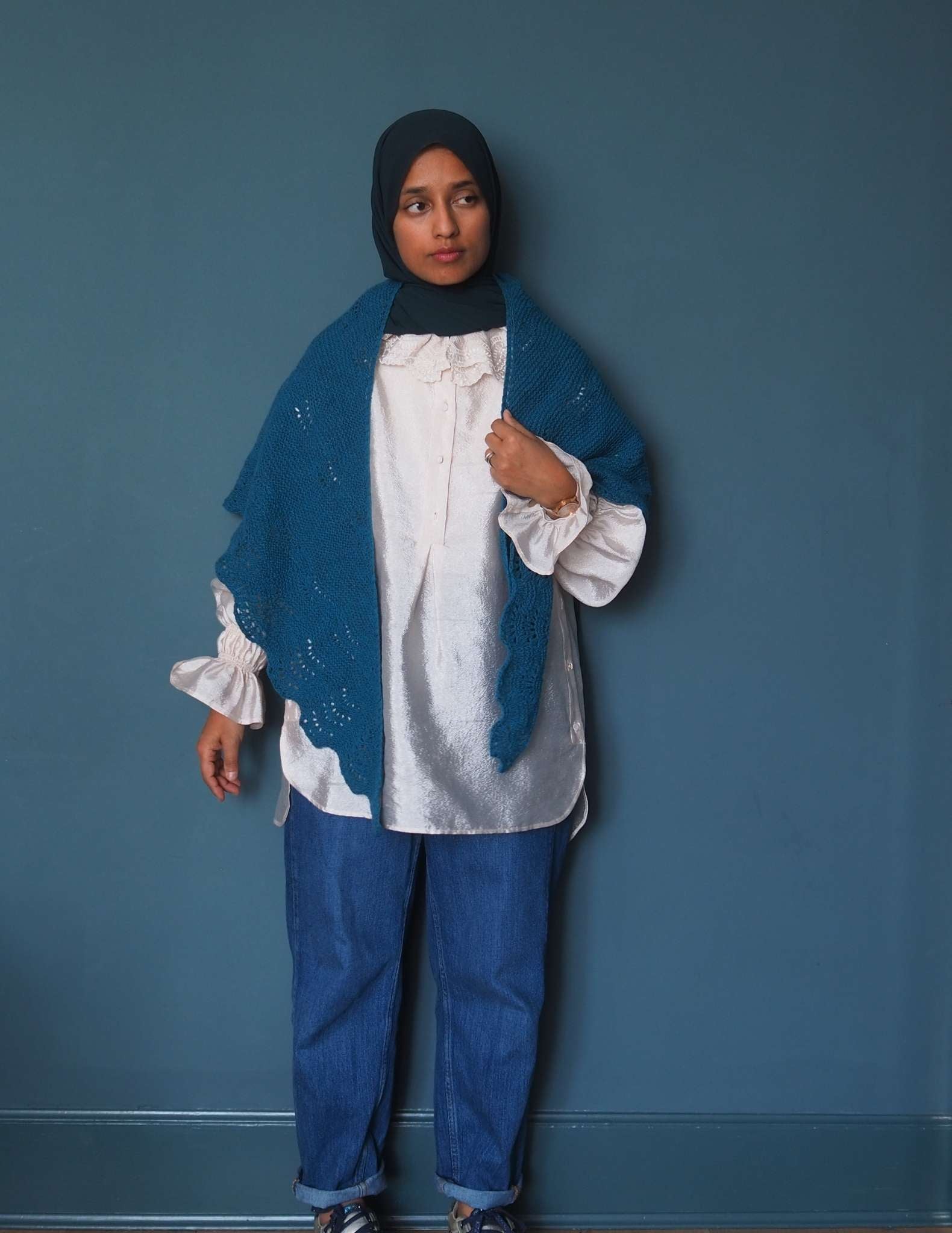 A brown woman in a hijab wears a ruffled white shirt and jeans, and stands in front of a dark blue background. She is wearing a blue wool shawl around her neck with the pointed ends hanging down in the front.