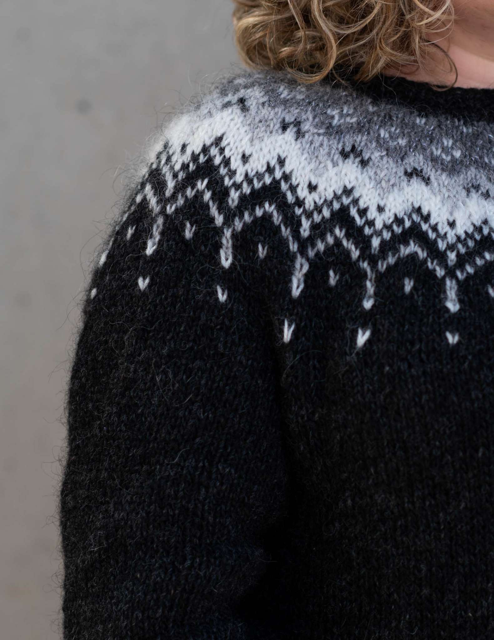 A close of the shoulder of a model with curly hair wearing a colourwork sweater in black with a contrast yoke in greys and white.