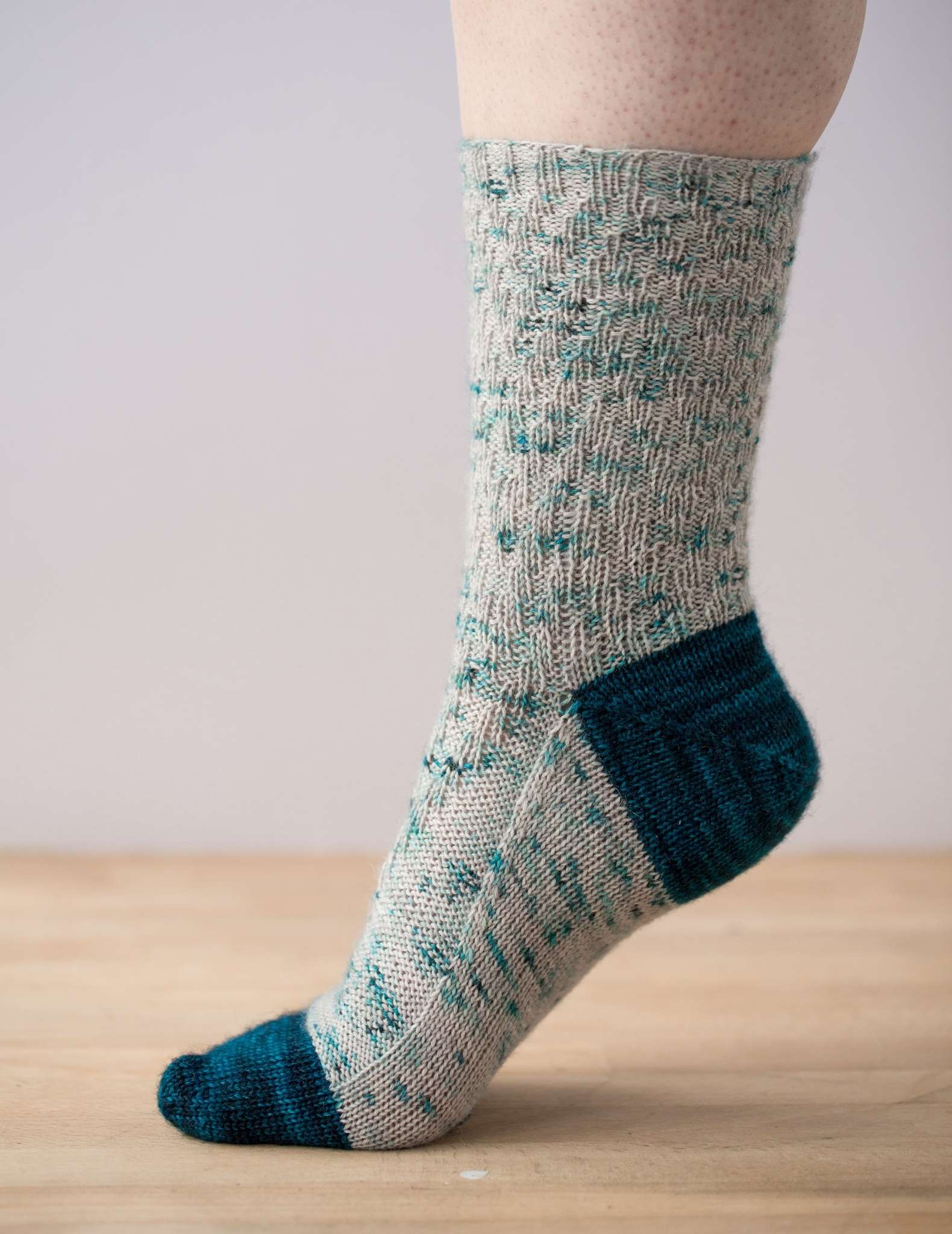 one foot is photographed from the side, with the toe on a wooden surface and the heel raised. The sock has a dark teal toe and heel and a paler textured pattern on the rest of the sock.