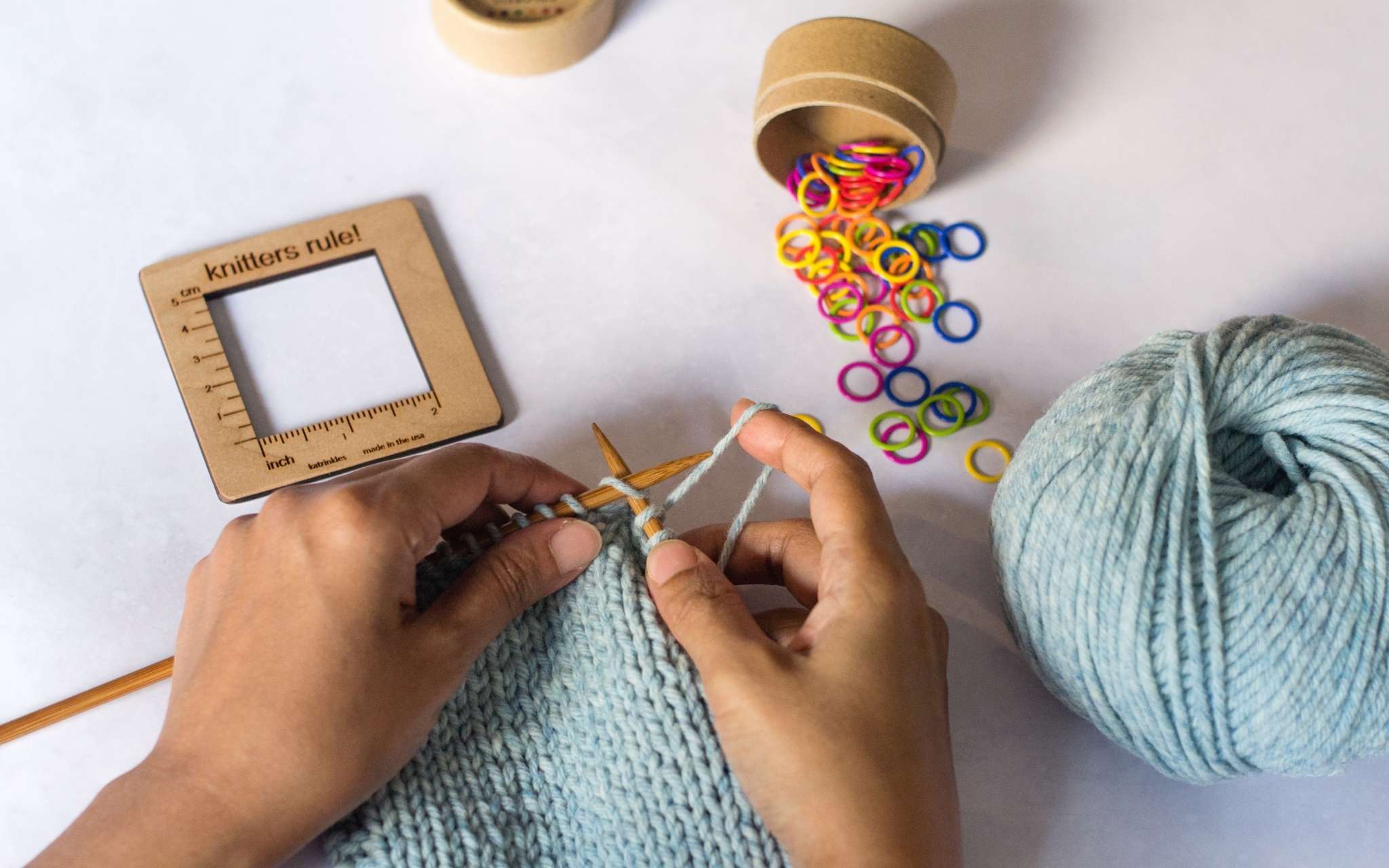 A woman knits on a pale blue swatch, her hands are visible from the bottom of the image and she has the yarn wrapped around her right finger as she knits. Above her on a flat surface lie a square wooden measuring tool and a brightly coloured stitch markers spilling out of a pot.