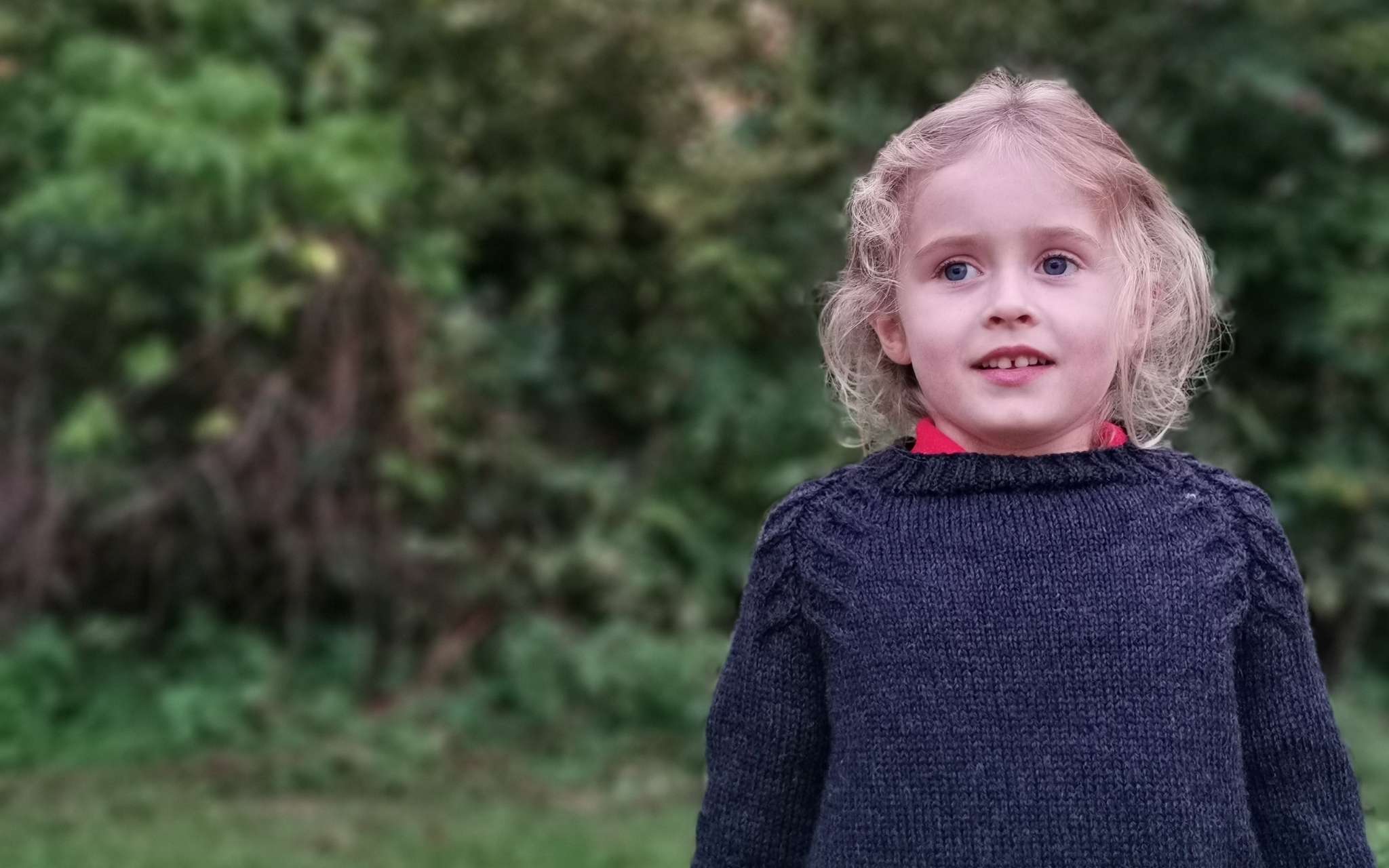 A young child with wavy blond hair wears a black sweater with cabled detailing along the raglan shaping. They are looking straight ahead and standing in a garden.