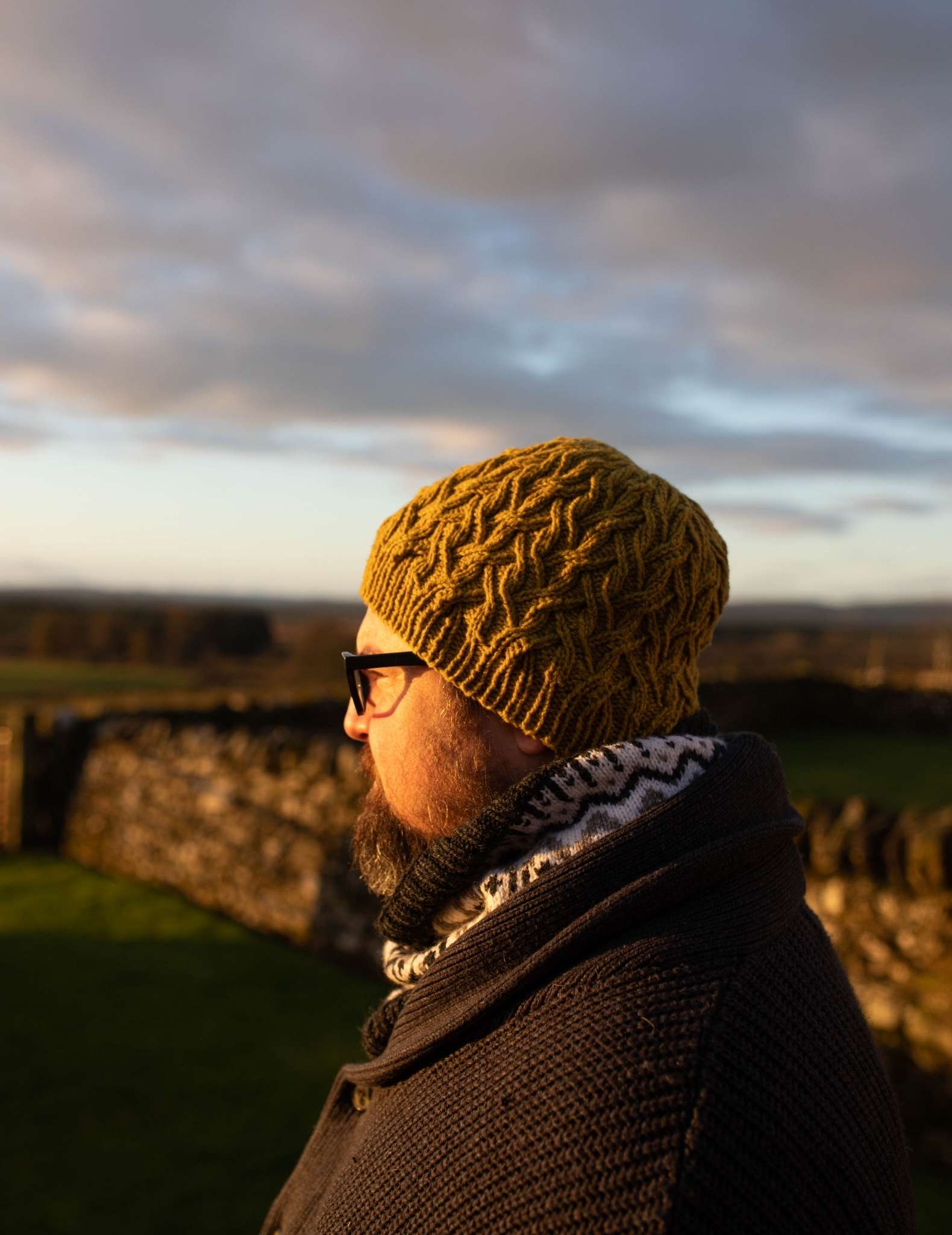 A white man with a beard and glasses looks out onto a rural landscape at sunset. He is wearing a green cabled hat and colourwork cowl
