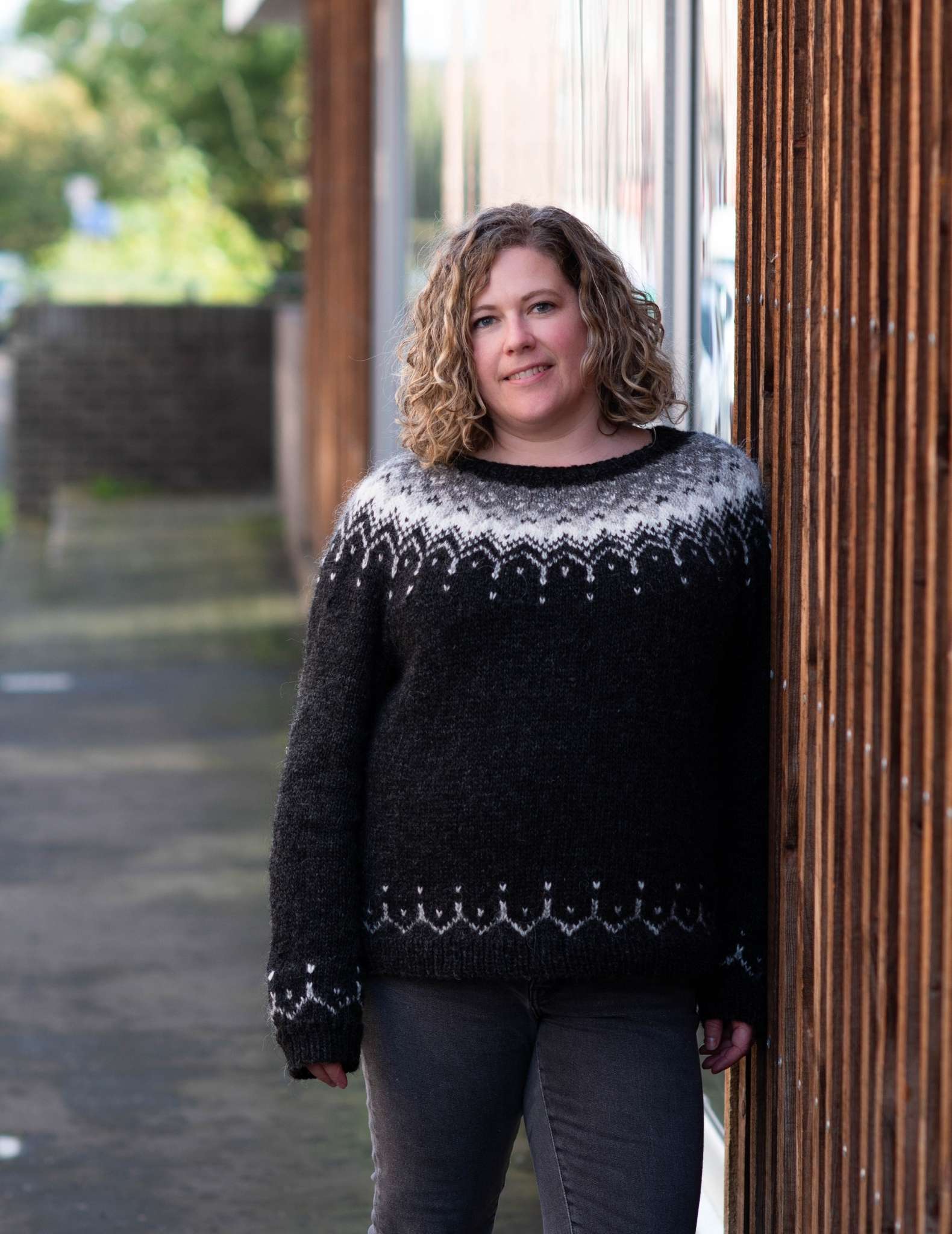 A white woman with curly hair wears a cosy black sweater with colourwork yoke in neutral colours, leaning against a wall