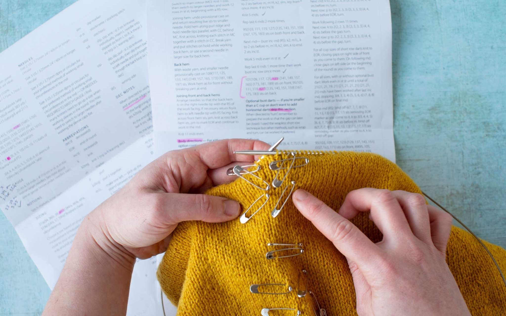 A yellow sweater marked with safety pins on the shaping points is held over a copy of a knitting pattern.
