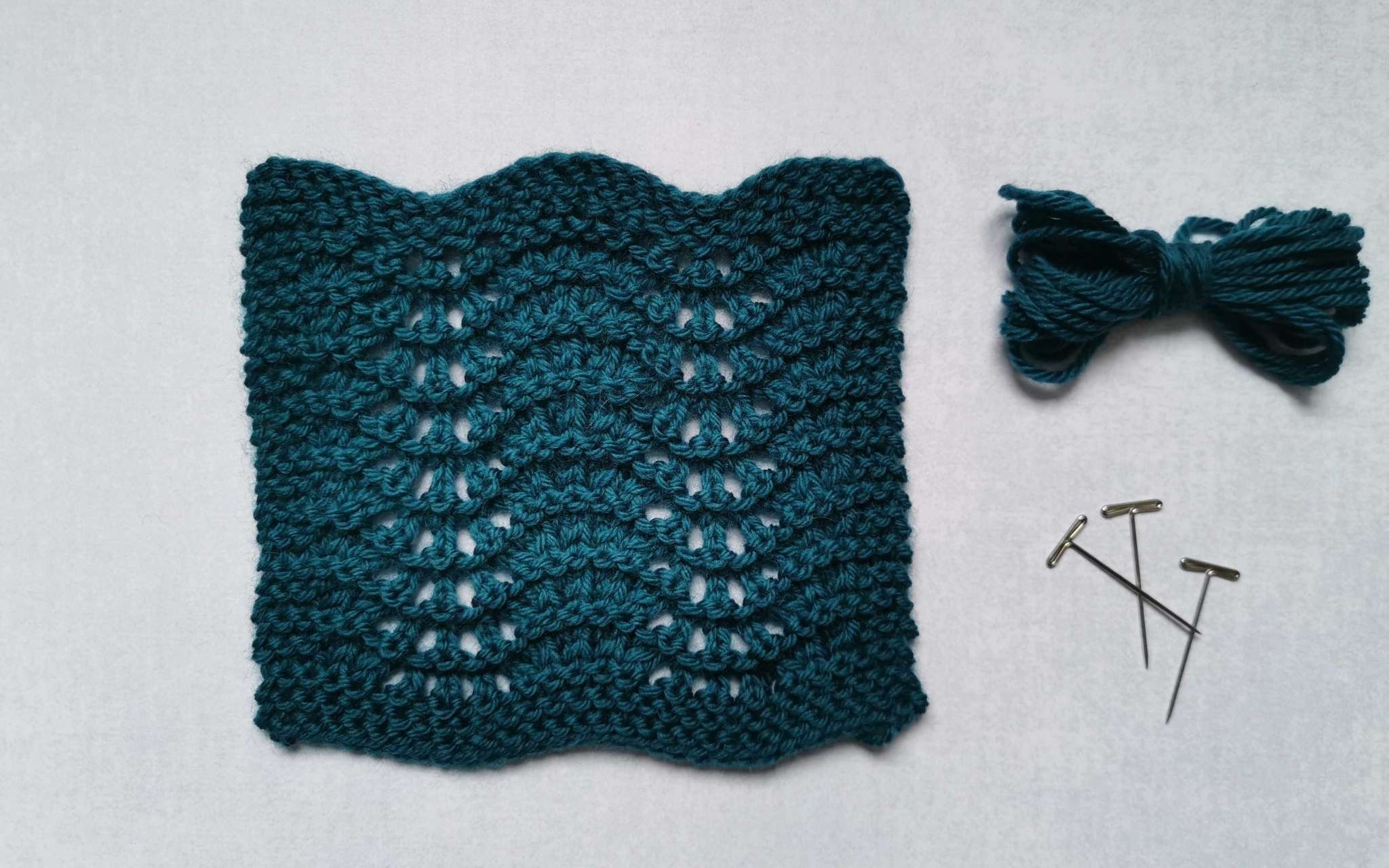 A square swatch of lace in teal, with some bundled up spare yarn and t-pins laying beside it to the right.
