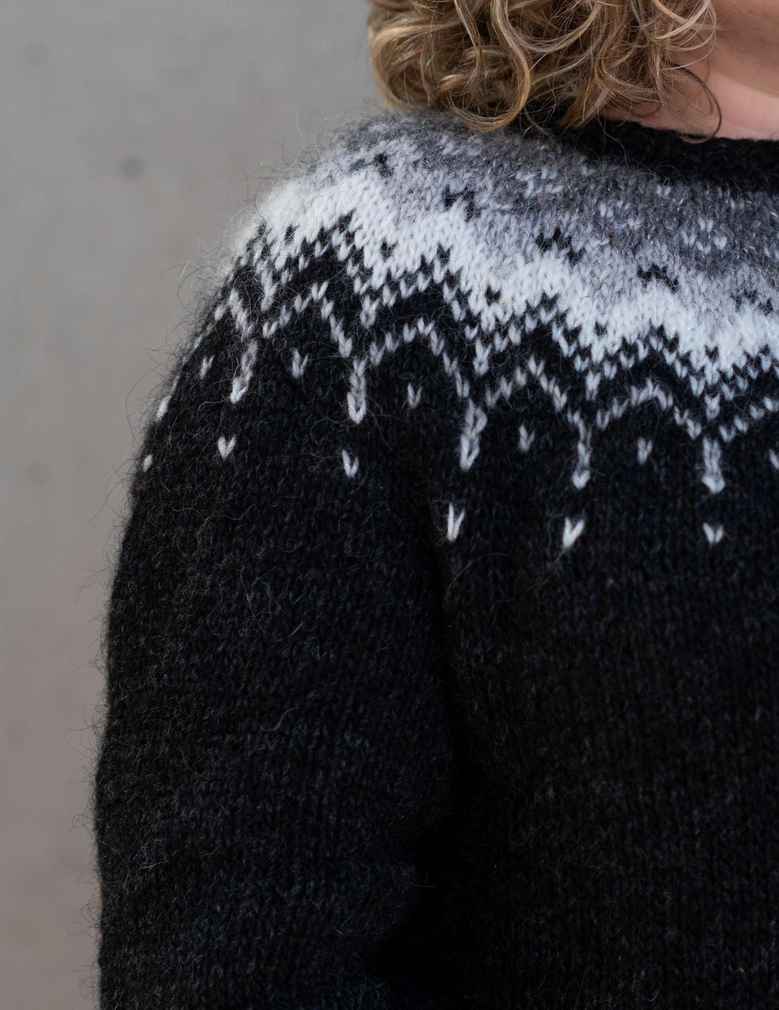 A close up of the shoulder of a white woman with curly hair wearing a dark sweater with colourwork yoke detail