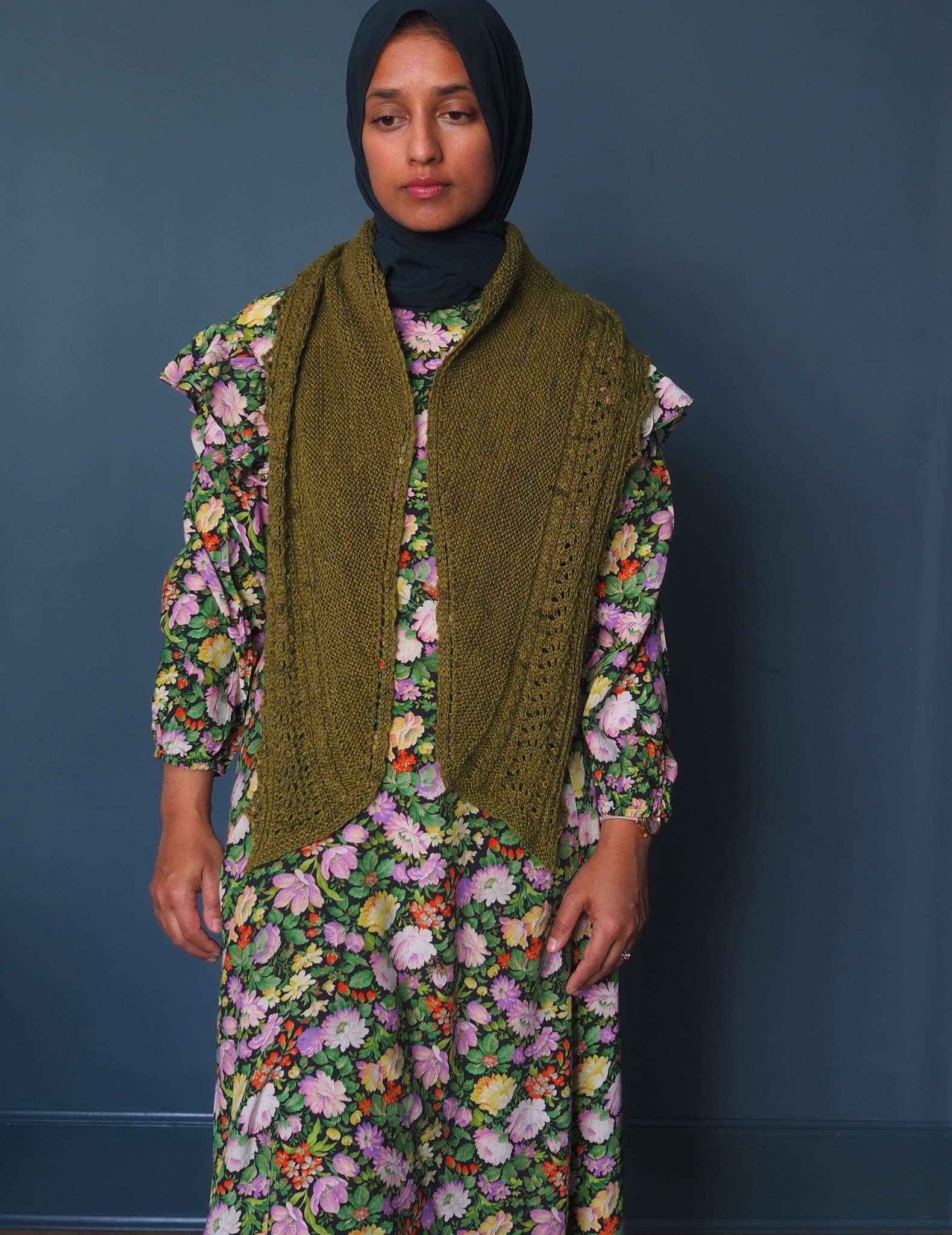 A brown woman in a hijab stands in front of a dark blue background. She is wearing a floral dress in shades of green, pink and yellow and a long green shawl is draped around her neck with the ends pointing down at the front.
