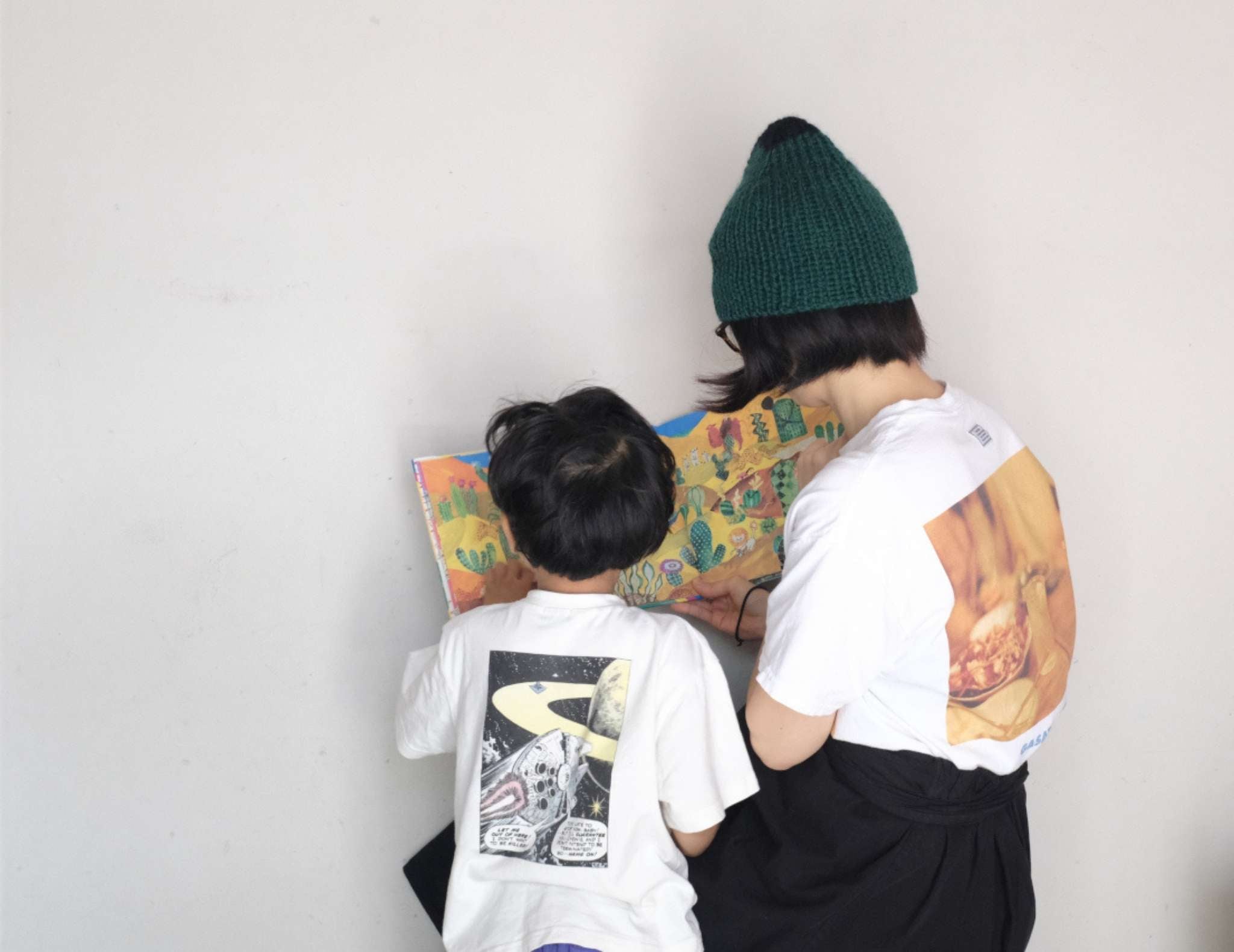 A small child and parent, both with straight black hair, are pictured from behind reading a picture book. They both wear white graphic t shirts and the adult has a teal ribbed hat.