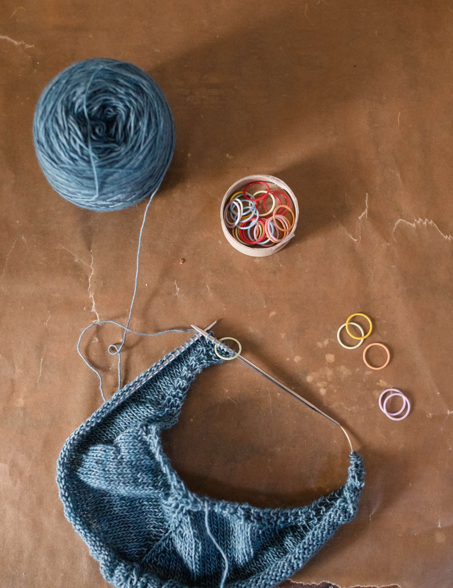 A flat lay image of a blue shawl still on the needles, a ball of attached blue yarn and pot of coloured ring stitch markers.
