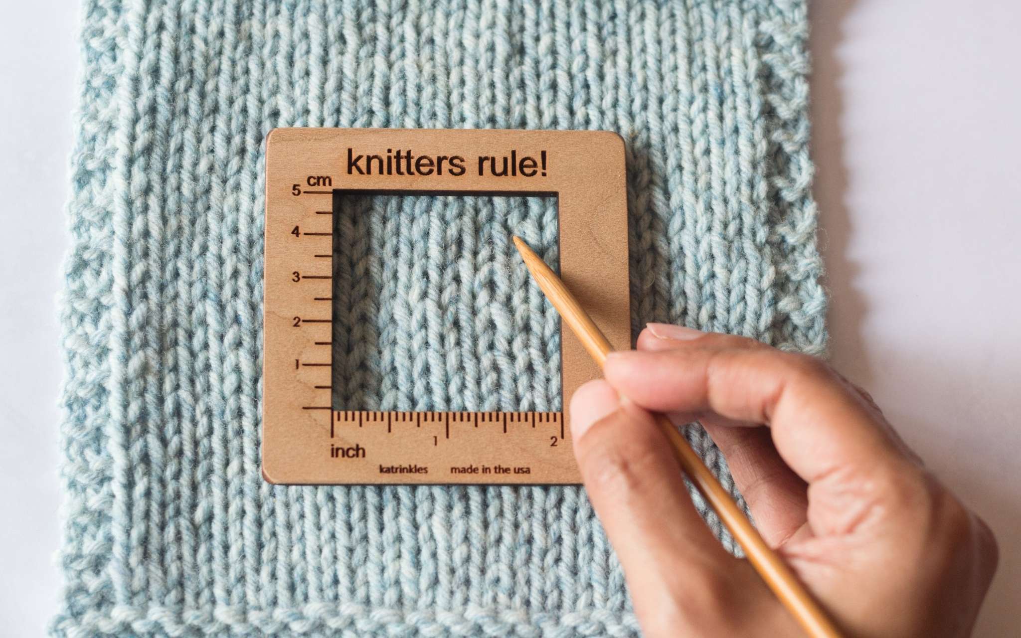 A pale blue swatch lays on a flat surface. A small square wooden measuring tools lies on top of the swatch and a wooden needle is being pointed towards the stitches in the centre.