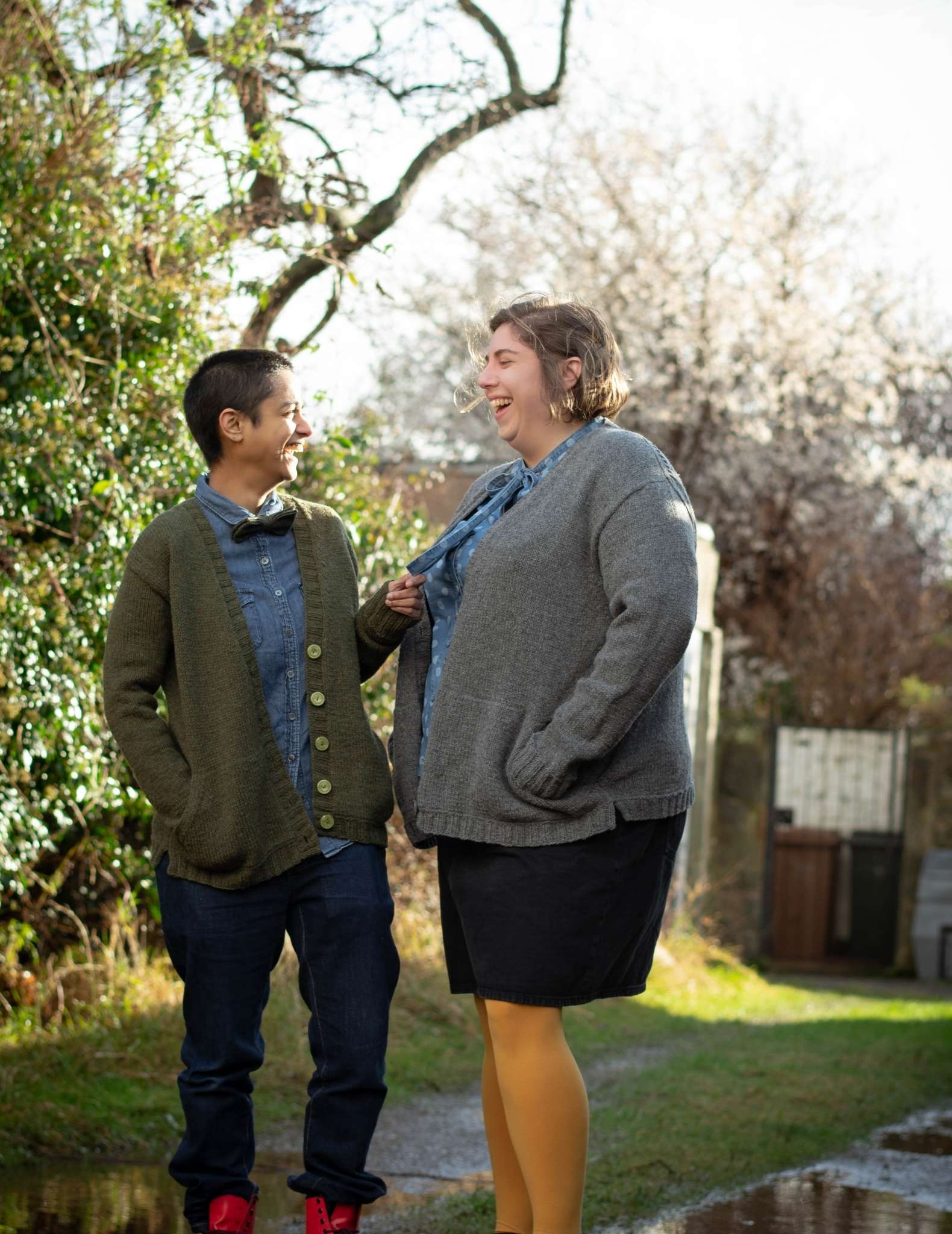 Two models wearing open, cardigans stand underneath some trees on a path through grass. They face each other and one holds the other's scarf, they are smiling at each other.