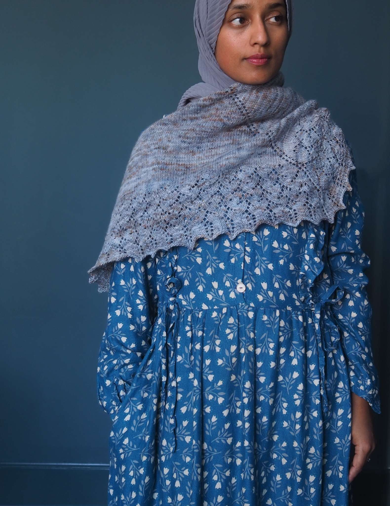 A brown woman in a hijab stands in front of a dark blue background and looks to the side. She is wearing a blue dress with small white floral print, and has a grey blue shawl wrapped around her shoulders and chest.