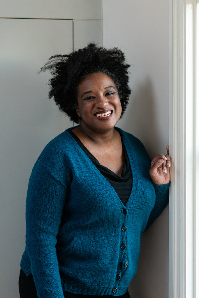 Woman with brown skin and dark hair laughing and wearing a dark top with a close fitting buttoned blue wardie 