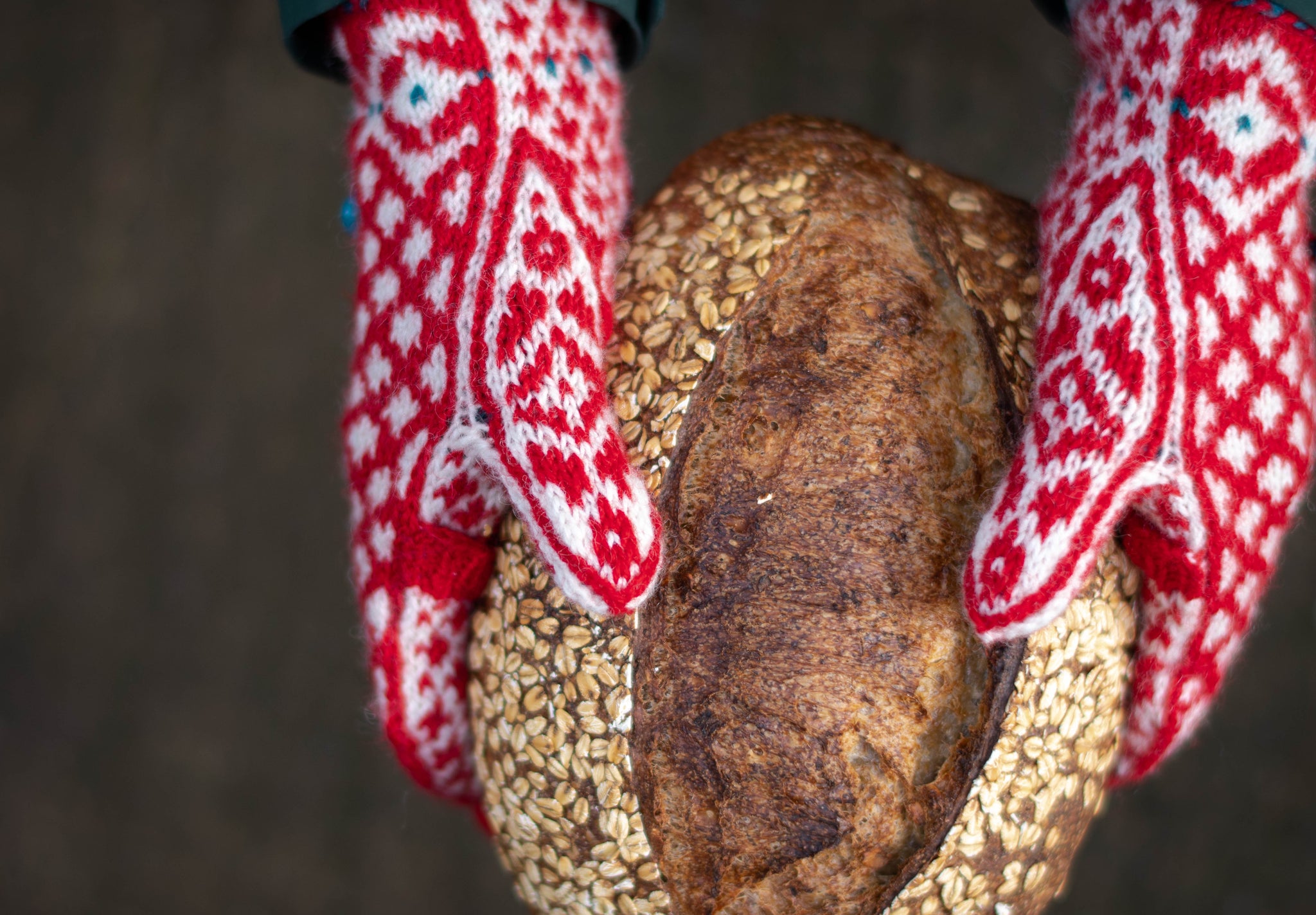 a pair of hands wearing read and white colourwork mittens hold a loaf of dark brown bread