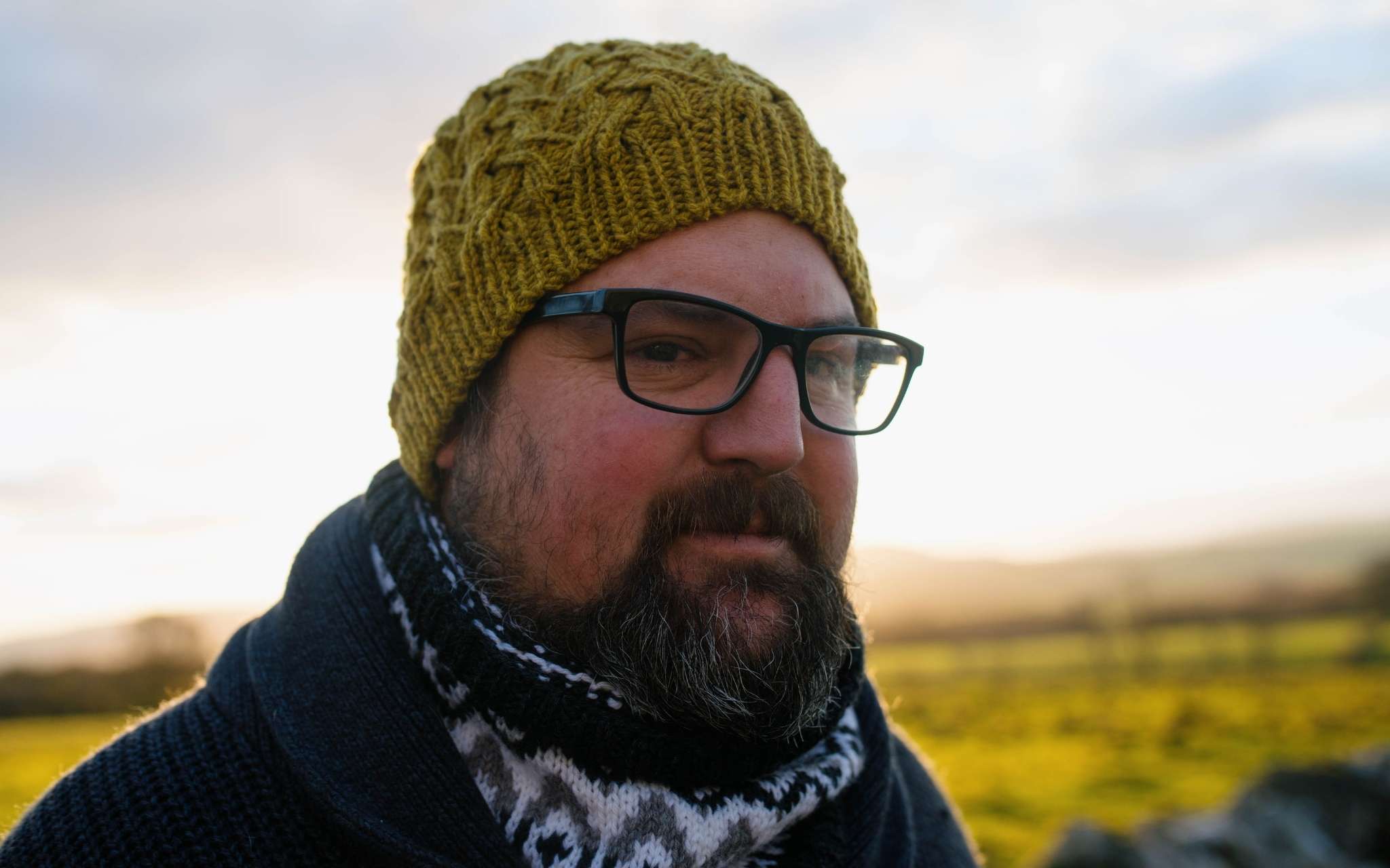 A white man with a beard and glasses stands in a rural landscape, looking to the right. He is wearing a green cabled beanie hat and colourwork cowl.
